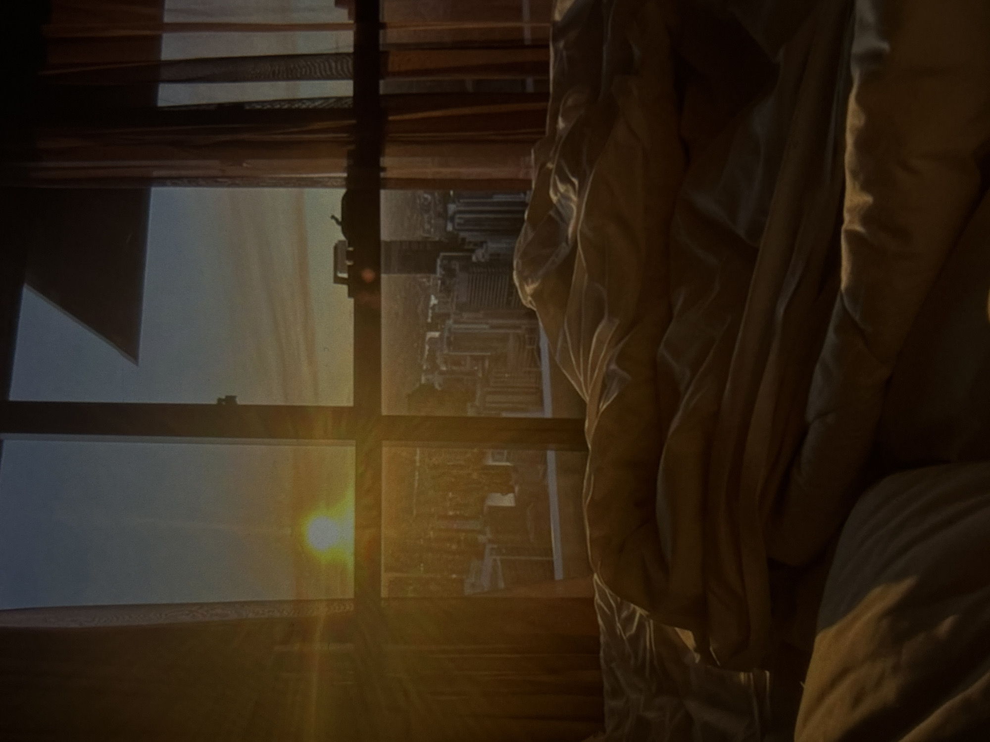 The image depicts a bed with rumpled white linens in the foreground. Beyond the bed, through a large window, a city skyline is visible, bathed in the warm glow of a sunrise or sunset. The sun, a bright, hazy orb, is positioned in the upper left quadrant of the window. The cityscape comprises numerous buildings of varying heights. Light orange curtains frame the window on both the left and right sides. The scene appears to be set in an urban environment, but no specific details indicate the location in Knezha, Bulgaria.