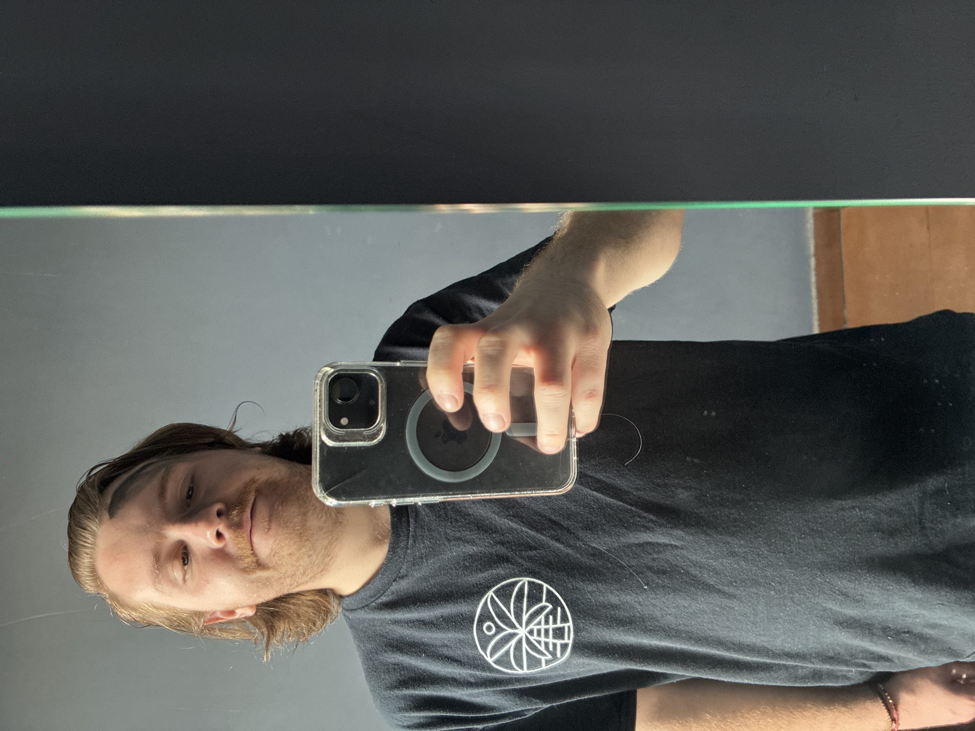 A man is taking a selfie in a mirror. He is wearing a black t-shirt with a white circular design on the left breast. He is holding a smartphone in a clear case with a MagSafe charger attached to the back. The background is a dark, possibly black, wall above the mirror. The environment appears to be indoors, likely a bathroom based on the presence of a mirror. The location in Cuautitlán Izcalli, Mexico, cannot be determined from this image.