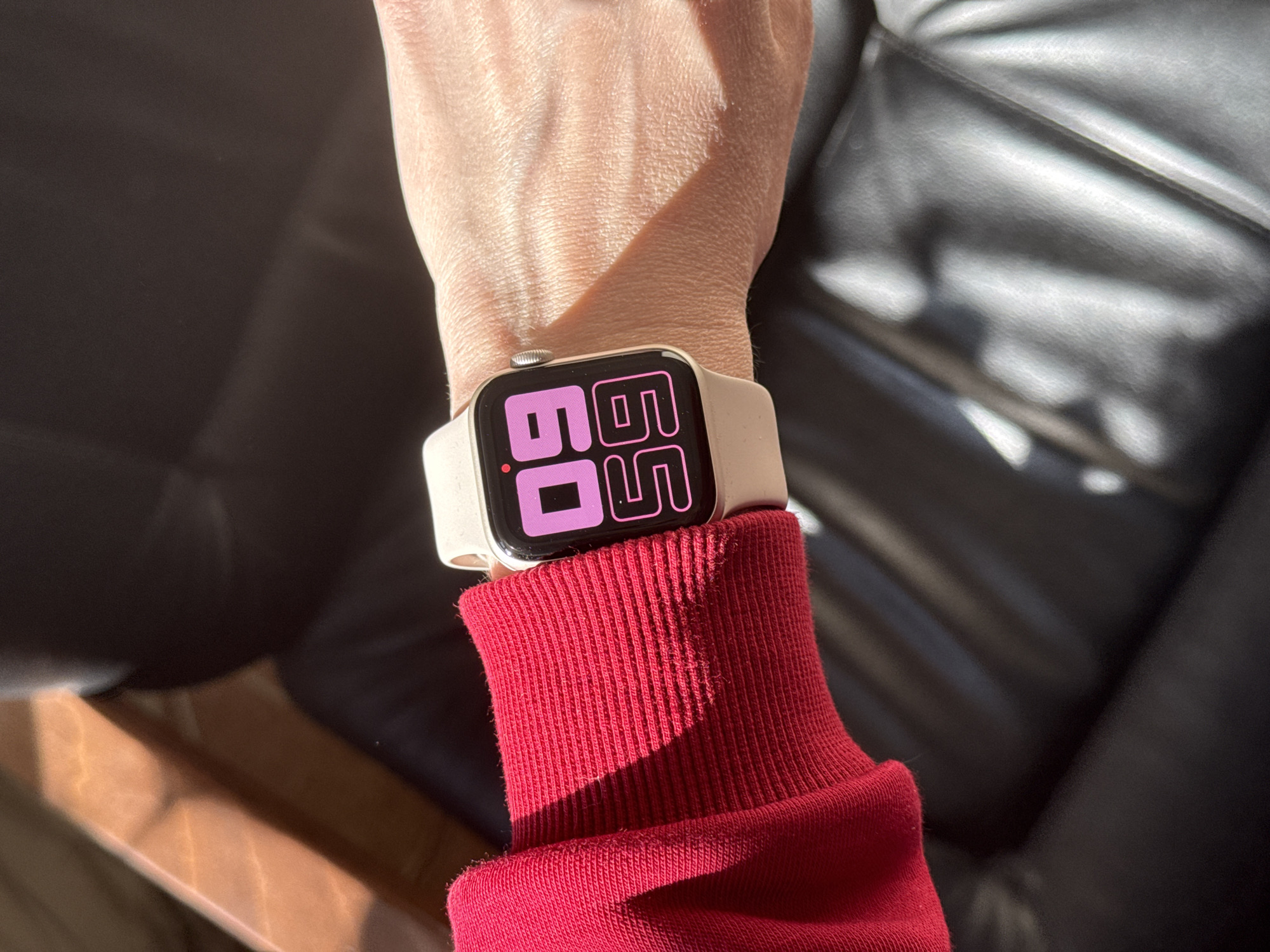 A close-up shot shows a wrist wearing a light-colored smartwatch displaying "60 55" in pink digits on a black screen. The wrist is fair-skinned and lies against a bright red, ribbed sweatshirt cuff. The background consists of a partially visible, dark-colored leather sofa and a wooden armrest, illuminated by natural light, casting a shadow across the wrist. The location is not discernible from the provided image.