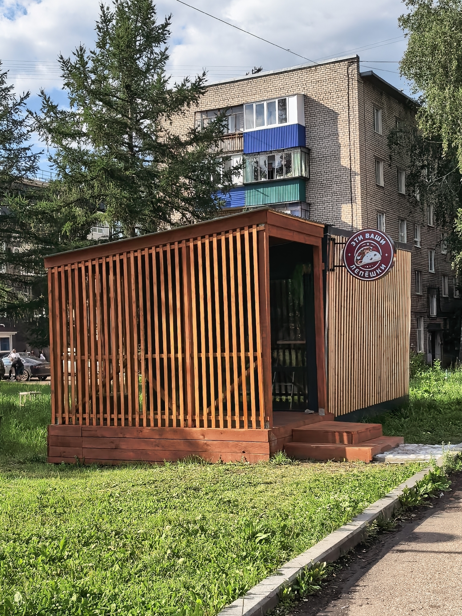 This media file depicts an outdoor dining area, likely a kiosk or small cafe, situated in a grassy area next to a sidewalk. The structure is made of wood, with a slatted wooden exterior and a wooden deck. It has an open front with steps leading up to it. A round sign with a red border hangs on the right side of the structure. Inside, there appear to be tables and chairs, suggesting it's a place for customers to eat.
In the background, a multi-story brick apartment building stands, featuring balconies with colorful accents of blue and green. Tall evergreen trees are also visible, one prominently in the foreground to the left of the building. The grass is green and well-maintained, with some weeds growing along the sidewalk's edge. A sidewalk and a road are also present.
On the left side of the image, a few people and cars are visible, indicating some pedestrian and vehicle traffic in the vicinity. The lighting suggests it is daytime, with the sun casting shadows, implying it could be late morning or afternoon. The sky is partly cloudy.
The text on the circular sign is in Russian and reads "ЭТИ ВАШИ ЛЕПЁШКИ" (ETI VASHI LYOPYOSHKI), which translates to "These are your flatbreads." This text, along with the image of a flatbread on the sign, clearly indicates the establishment serves flatbreads or similar food items. The overall atmosphere appears to be casual and functional, set in a typical urban residential environment.