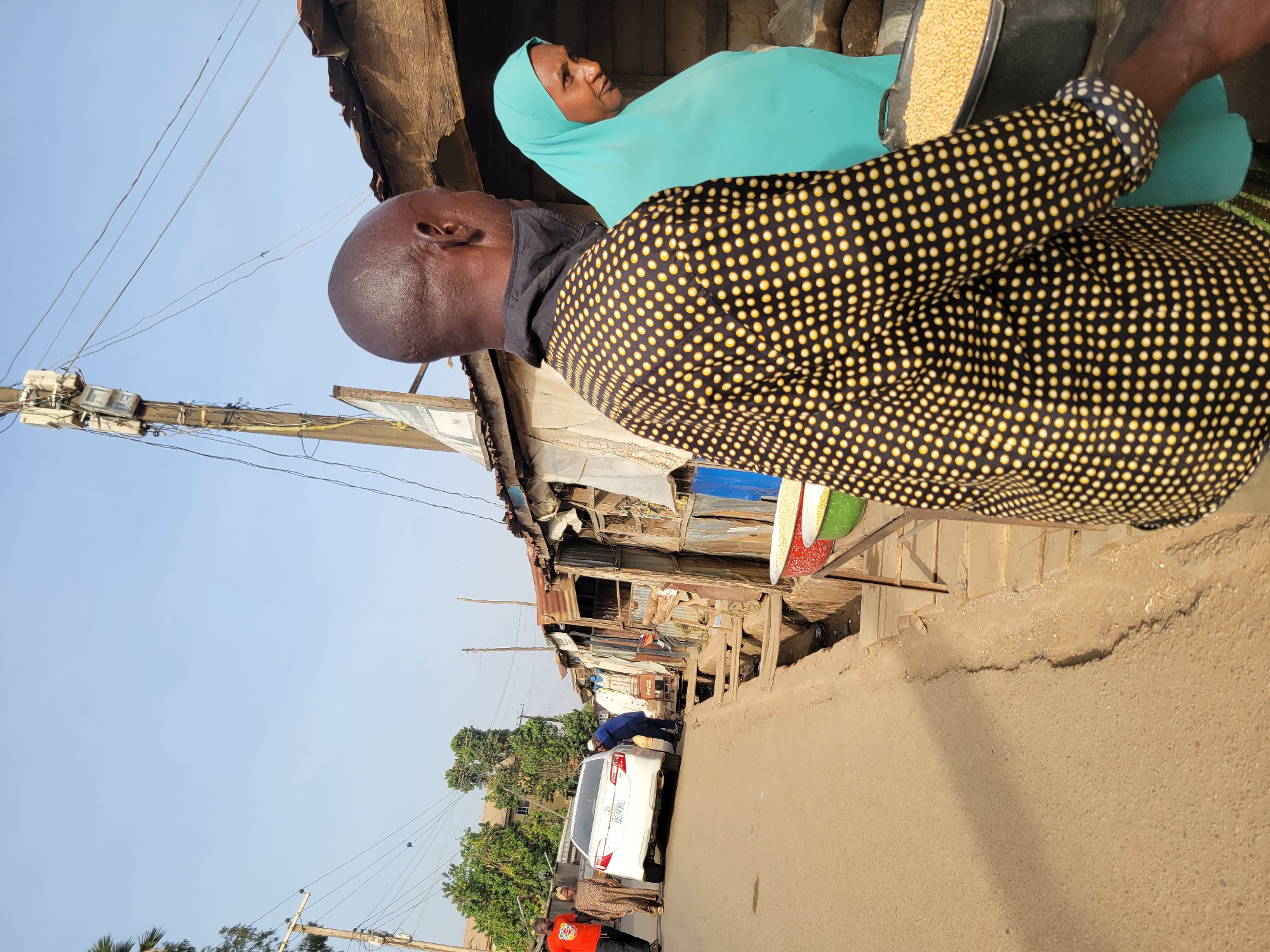 The scene shows an outdoor street view in Jos, Nigeria. A man with a shaved head and wearing a black shirt covered in small yellow dots stands next to a woman wearing a light blue hijab. The woman has her head turned towards the man. The scene appears to be a marketplace, with various makeshift stalls visible in the background. A white car is parked in the street. Other individuals are standing near the car and in the background. Power lines run overhead. The sky is clear and light blue. The street is made of sand.