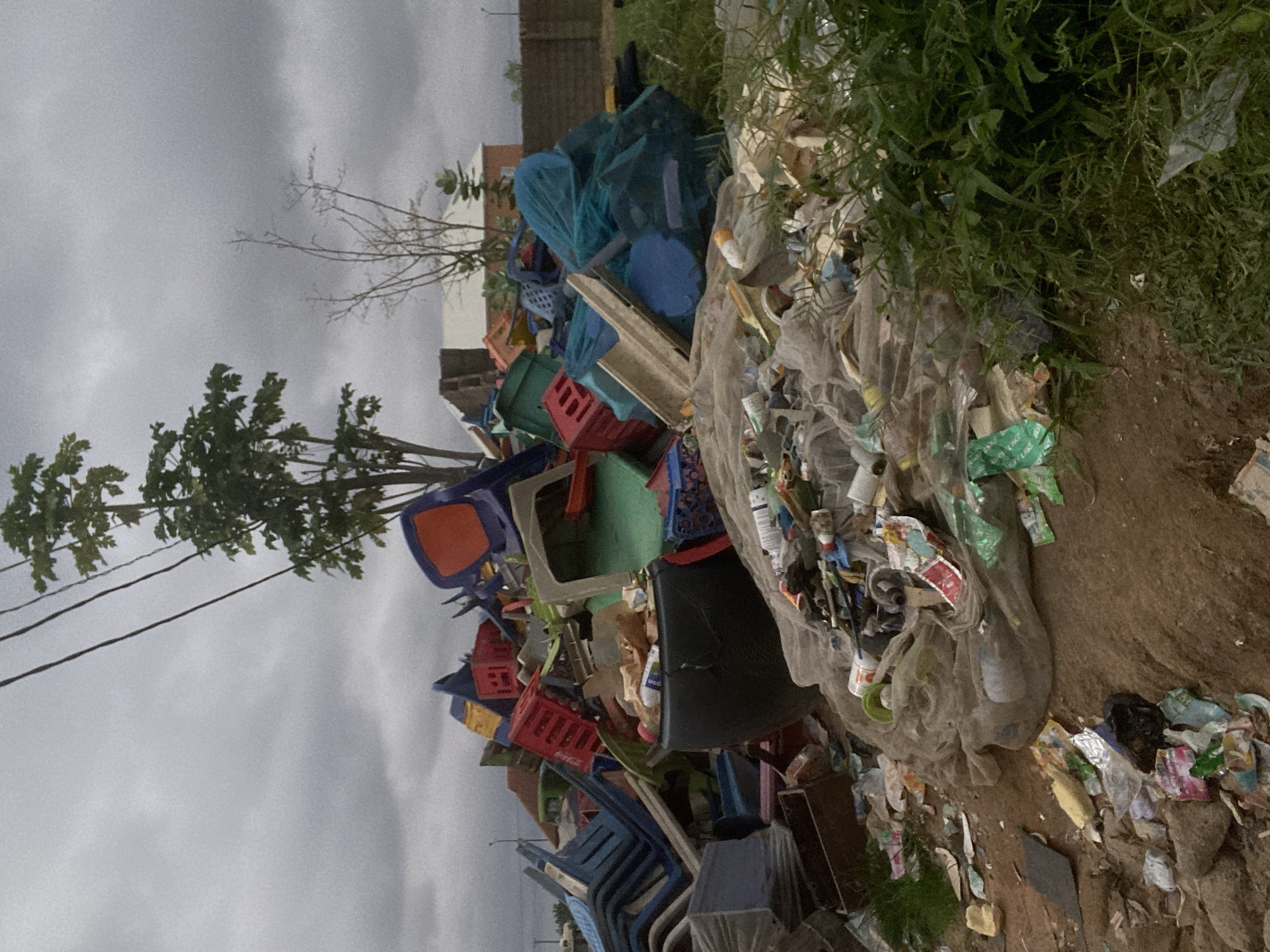 A large, informal dump site is visible in Gbaza, Nigeria, under an overcast sky. The foreground is dominated by a large pile of assorted plastic waste, including discarded chairs, crates, and various containers. A prominent black bucket or tub is situated in the mid-ground, filled with debris. In the background, a tree stands tall amidst the refuse, with electrical wires overhead. The scene suggests a significant accumulation of waste, characteristic of an unregulated disposal area. The surrounding environment appears to be a mixture of dirt and overgrown vegetation.
