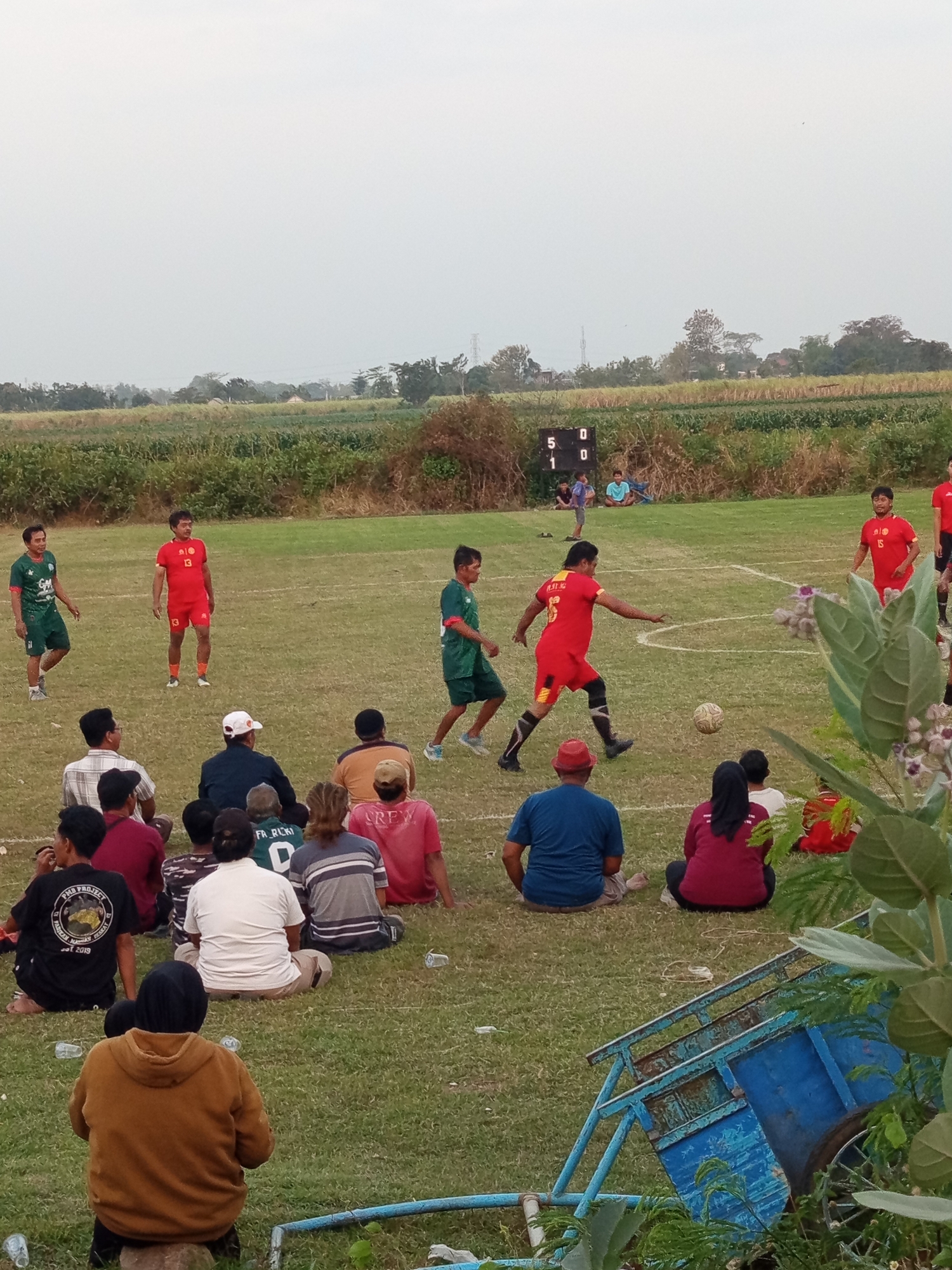 The image depicts an outdoor soccer match taking place on a grassy field in Mrican, Indonesia. In the foreground, spectators are seated on the grass, some facing away from the camera, while others are partially visible. A blue, metal cart or trailer is positioned in the lower right quadrant of the frame. In the midground, two soccer players in red uniforms are actively engaged in the game, with one kicking a soccer ball. Other players in green and red uniforms are scattered across the field. A scoreboard is visible in the background, displaying the score "5 0" and "1 0". The surrounding environment consists of a grassy field with agricultural land and trees in the distance, under a hazy sky.