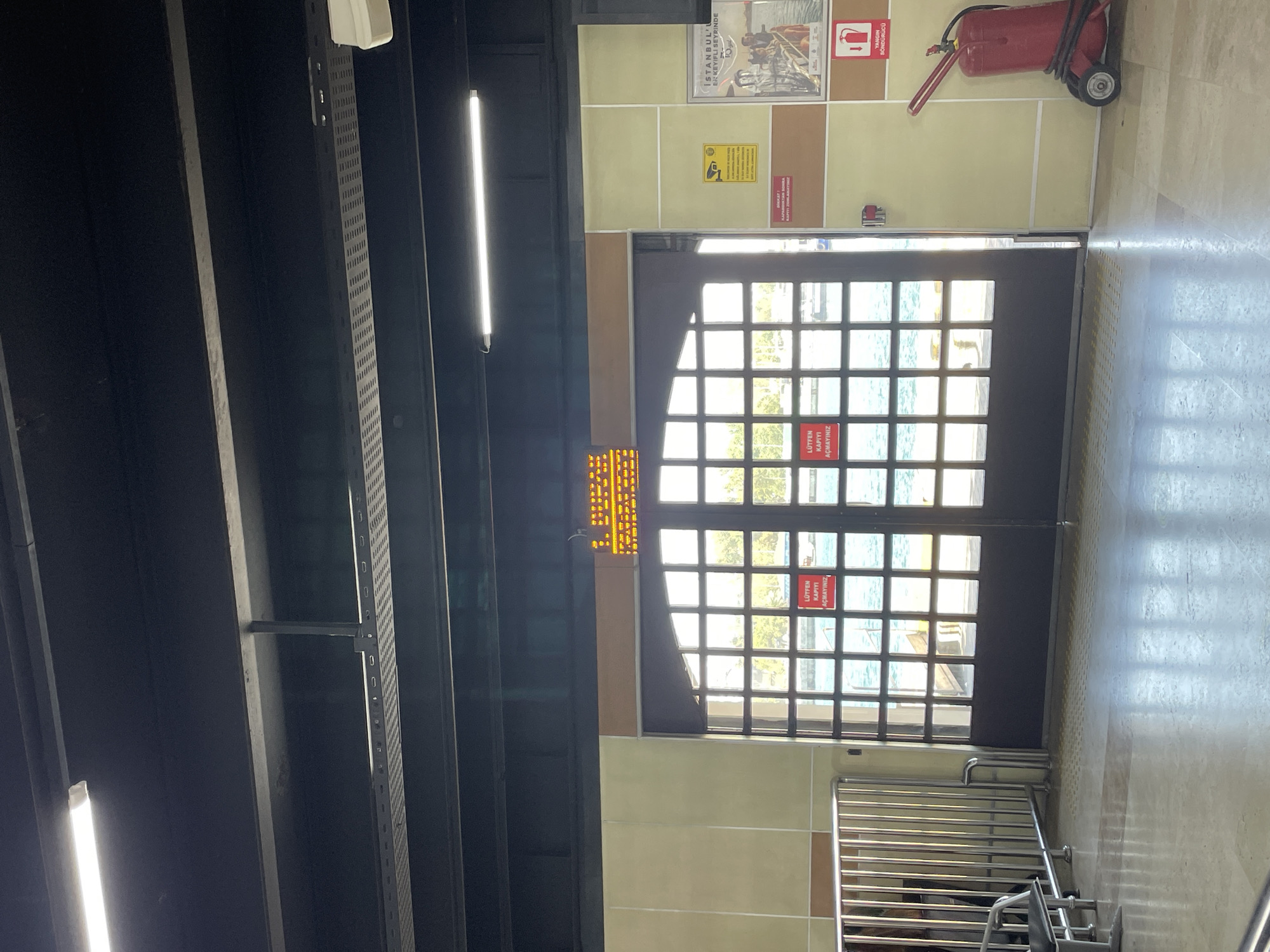 The image shows a view from inside a building, looking out through a set of large glass double doors towards a body of water. The doors are framed by dark wood and are divided into a grid of square panes, through which sunlight reflects off the water. Outside, trees and a boat are visible.
On the left side of the image, a metal barrier and a bench are present. On the walls, various signs are displayed. A prominent yellow sign with a camera icon and text is on the right wall, accompanied by a poster titled "ISTANBUL'ER KEYİFLİ SEYRİNDE". Below these, a red fire extinguisher on wheels is positioned. On the doors themselves, red signs with white text read "LÜTFEN KAPİYİ AÇMAYINIZ". An electronic display board with orange text is mounted above the doors on the interior wall. The floor is tiled and reflects the light. The ceiling is dark with exposed lighting fixtures. This scene appears to be a ferry terminal or a similar transit point in Kadıköy, Turkey, likely near the Bosphorus Strait given the maritime view.