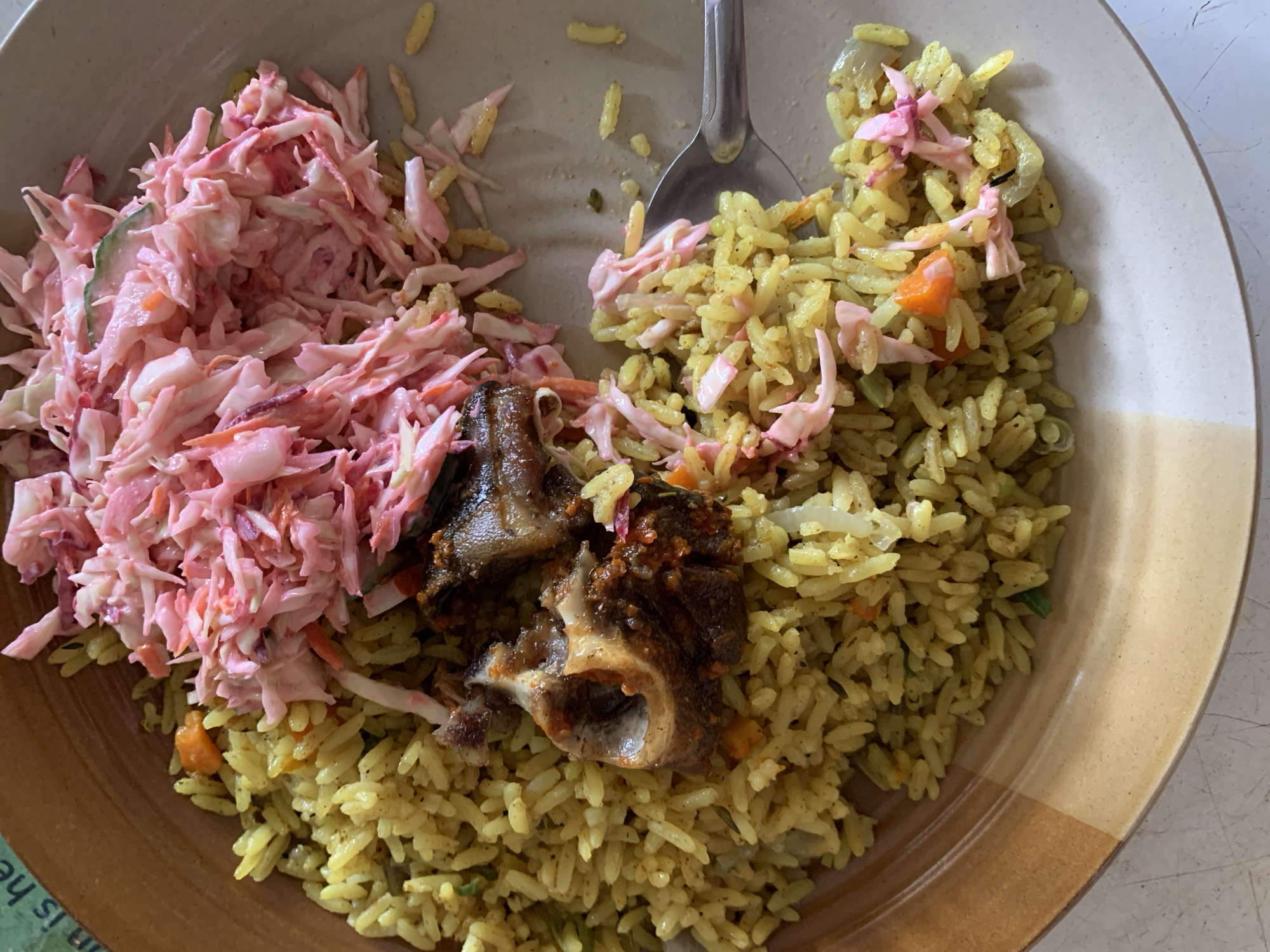 A plate of food is viewed from directly above. The plate is beige with a brown rim. The food consists of yellow rice, a pink coleslaw, and two pieces of what appears to be cooked oxtail. A metal spoon is visible partially buried in the rice. The meal is on a flat white surface. The plate may be located near the New Stadium, Nigeria, given the request.