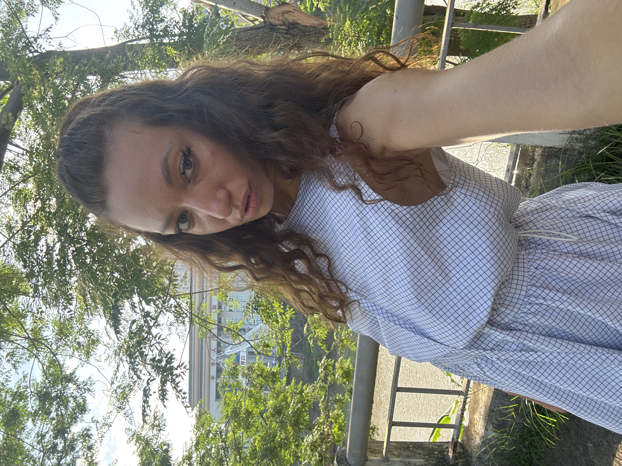 A woman is taking a selfie outdoors, positioned next to a metal railing. She is wearing a light blue and white checkered dress. Her hair is long and wavy. The background shows green foliage, a glimpse of a building, and part of a city scene, suggesting the photograph was taken in a semi-urban environment. The building visible in the distance features Chinese characters, which corroborates the location in Lao Cai Ward, Vietnam, given its proximity to the Chinese border. The lighting is bright, suggesting it is daytime.