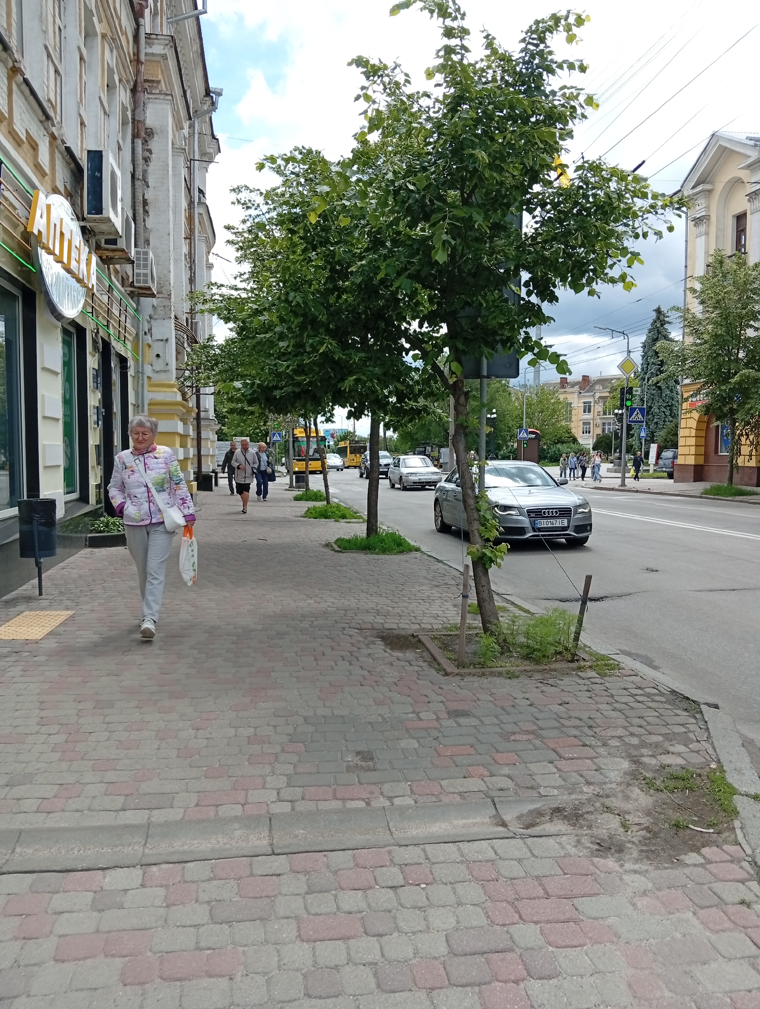 A medium, eye-level shot shows a city street scene in Poltava, Ukraine. An elderly woman, wearing a light floral-patterned jacket and gray pants, walks on a brick sidewalk towards the viewer on the left side of the frame, carrying a white bag. Behind her, two people walk in the same direction, while a bus and other cars are visible further down the street. The street is bordered by buildings on both sides, including a pharmacy with a large, round sign written in Cyrillic. In the background, several people are crossing a street at a marked crosswalk. Trees line the sidewalk on both sides of the street, partially obscuring some of the buildings and vehicles. A silver Audi is parked on the right side of the road. Overhead power lines are present. The sky is overcast with some hints of blue.