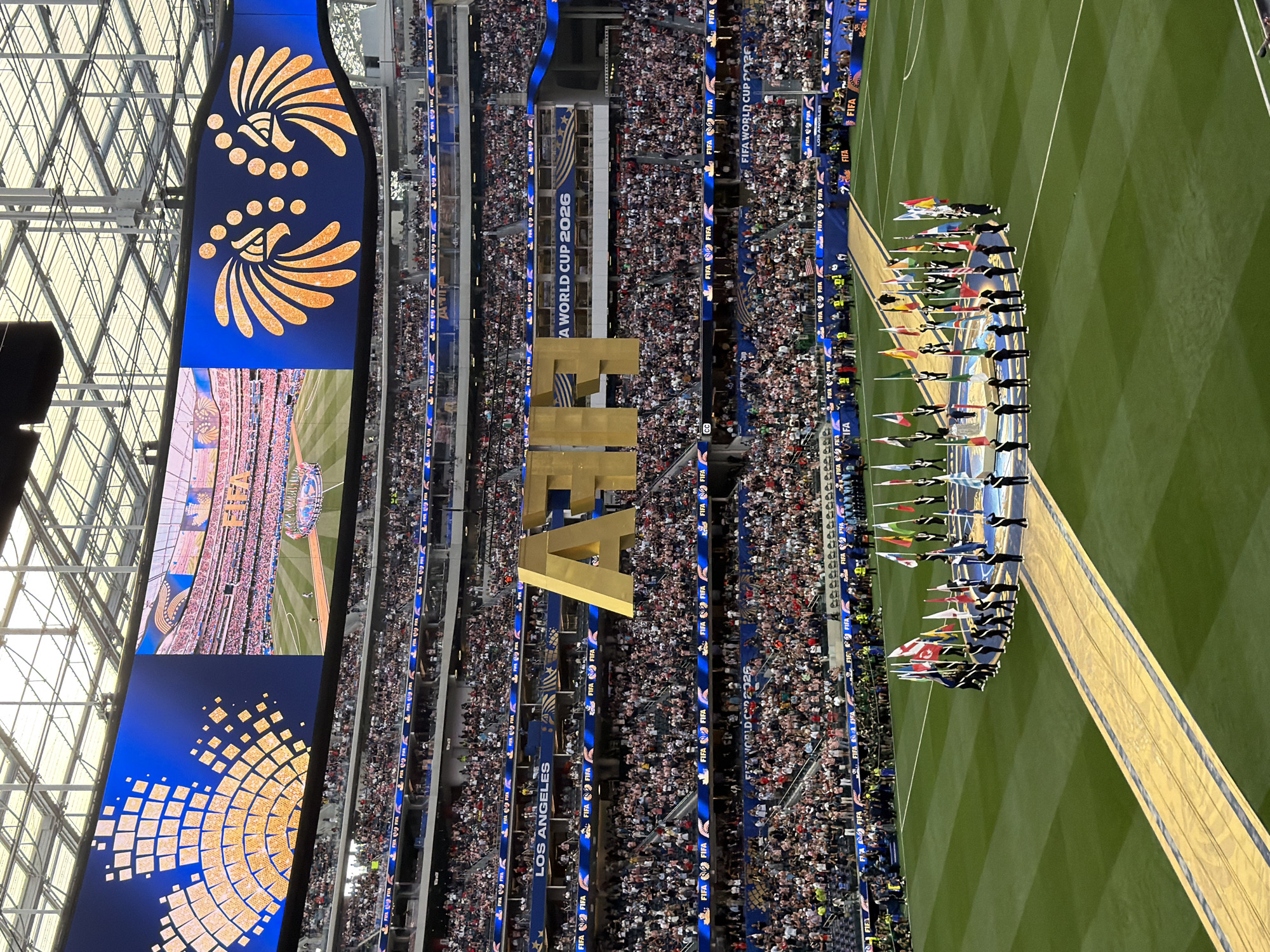 The image captures a wide view of an outdoor stadium filled with a large crowd of spectators, likely for a sporting event. In the foreground, a green soccer field is visible, with a golden pathway leading towards a semi-circle formation of individuals holding various national flags. The people in the formation are dressed in dark uniforms, and some are holding what appear to be trophies.
Dominating the center of the image, partially obscuring the field and the crowd behind, are large, golden three-dimensional letters spelling out "FIFA". Above the field, a massive video screen displays content, with the left side showing a blue background with a geometric orange design and the right side showing a packed stadium and the "FIFA" logo, along with text reading "A WORLD CUP 2026". Above this screen, the stadium's roof structure is visible, with lights and infrastructure.
The stands are packed with a diverse crowd, many of whom are looking towards the field. Along the upper tiers of the stadium, banners and digital displays featuring "FIFA" branding and "LOS ANGELES" are visible. The overall atmosphere suggests a significant event, possibly an opening ceremony or a key match. The location is identified as Inglewood, United States of America, a city known for hosting major sporting and entertainment events in its stadiums.