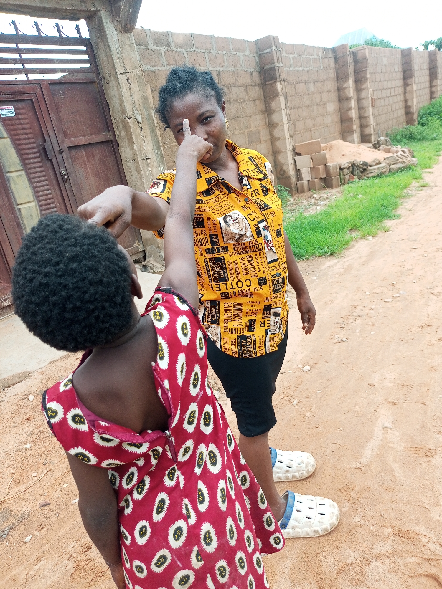 In a sandy, outdoor environment in Tempure, Nigeria, a woman in a yellow, patterned shirt and black pants points her finger towards the head of a child wearing a red, patterned dress. The child is facing away from the camera, and the woman is looking directly at the camera with her finger extended. In the background, a brick wall and a metal gate are visible. The ground is composed of reddish-brown sand and some sparse green vegetation.