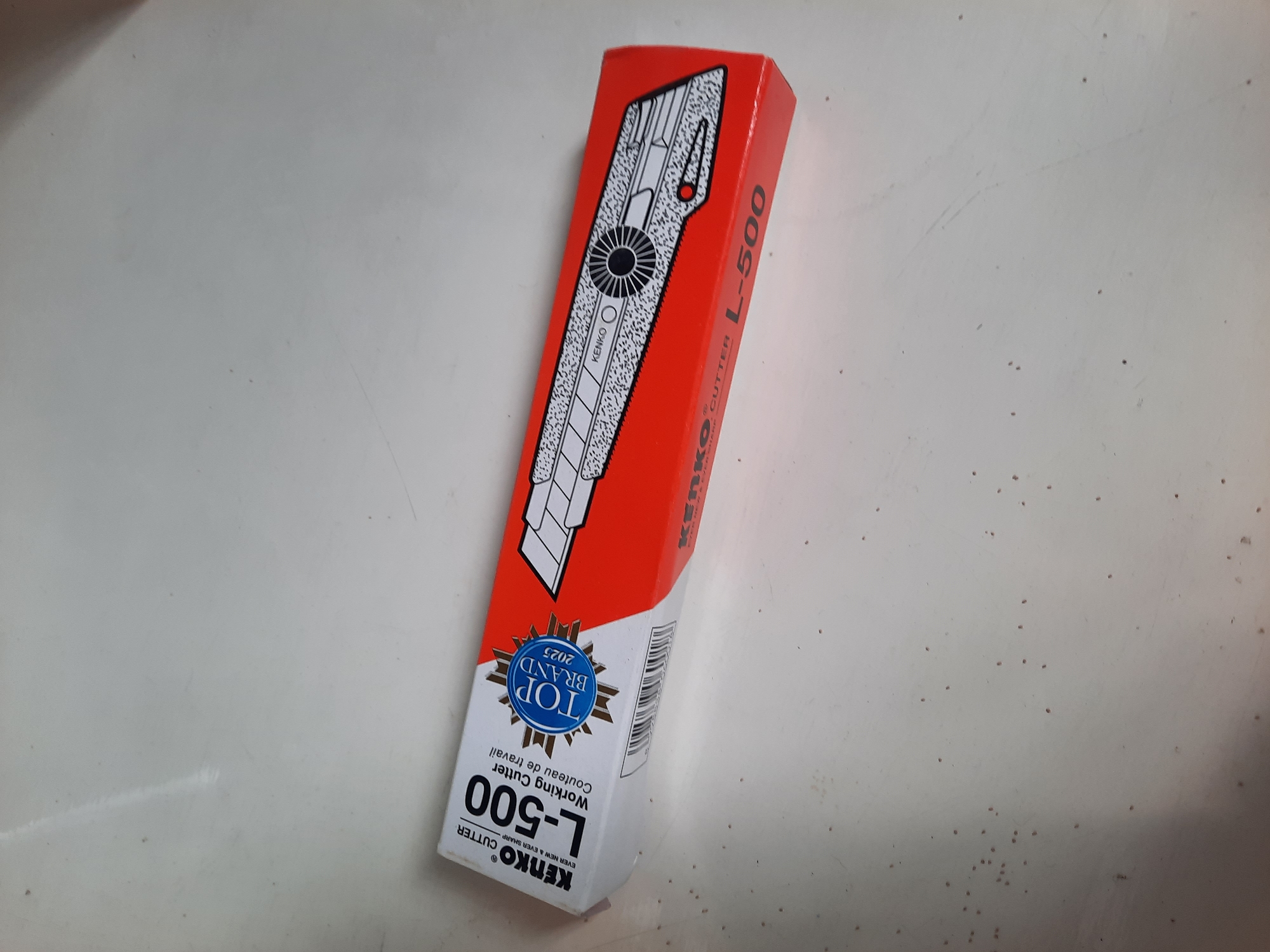 A long, rectangular box for a KENKO L-500 Working Cutter is angled on a light-colored, possibly white, surface. The box is primarily orange and white, with the brand name "KENKO" and the model number "L-500" prominently displayed in large font. A graphic of a utility knife is printed on the orange side of the box. The text "TOP BRAND 2025" is within a blue starburst logo on the white side. The surface appears to be a tabletop or counter and has numerous small, dark specks scattered across it. The lighting suggests an indoor environment, likely a domestic or retail setting. There are no people or animals visible in the image. The location is not identifiable from the visual information provided.
