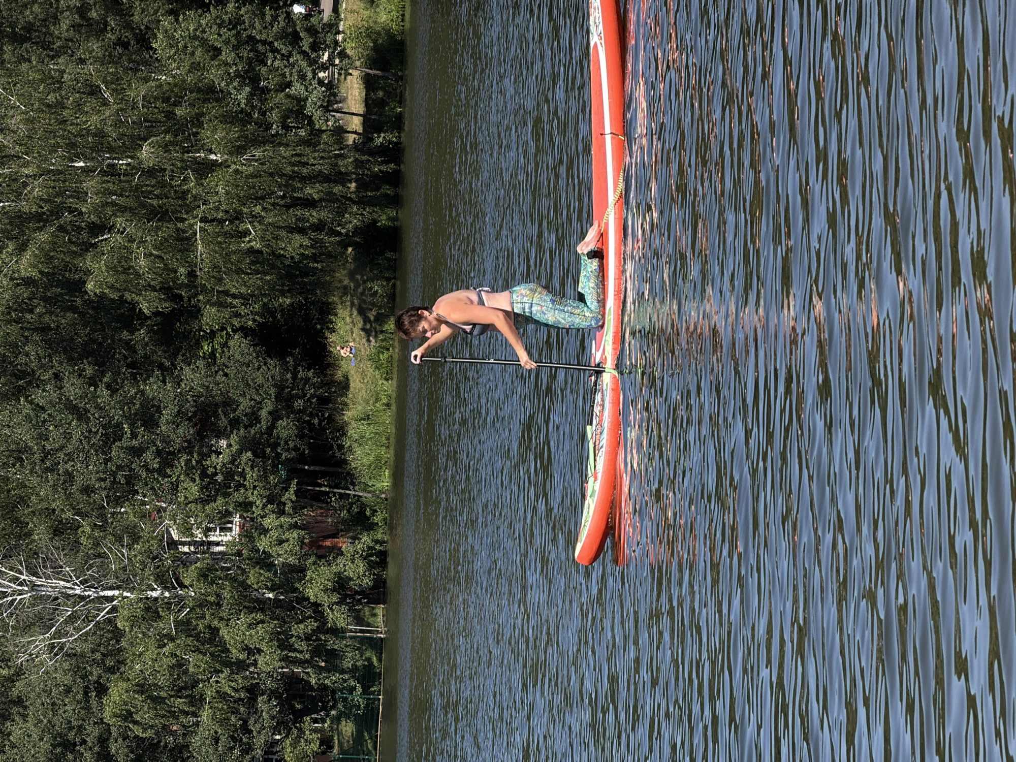 In Malakhovka, Russia, a woman kneels on an orange paddleboard in a body of water. She is wearing a patterned swimsuit and is holding a paddle. The paddleboard is positioned on the surface of the water, which is rippled and reflects the sunlight. In the background, there is a dense line of trees along the shoreline, with some buildings partially visible through the foliage. The sky is clear and blue, suggesting a sunny day.