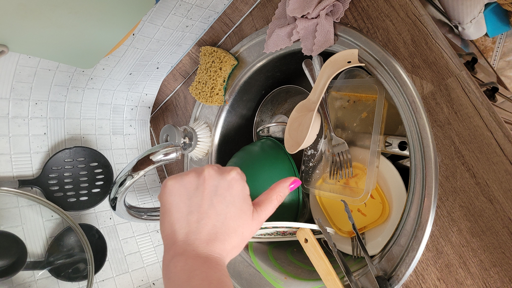 This is a high-angle, close-up shot of a cluttered kitchen sink filled with dirty dishes. The sink is made of stainless steel and contains a variety of items, including a yellow sponge, a green bowl, a wooden spoon, a fork, a clear plastic container with leftover food, and various plates and pots. A dishrag, appearing to be pink or mauve, is draped over the edge of the sink. To the left of the sink, a black slotted spoon and a pot lid are visible on a white tiled countertop. A chrome faucet with a dish brush attached is also present.
In the foreground, a person's left hand with bright pink nail polish is visible, giving a thumbs-up gesture. The hand appears to be positioned near the edge of the sink. The background shows a wooden countertop and the edge of a white tiled backsplash.
The scene suggests a domestic setting, likely a kitchen, where dishes are piled up, possibly after a meal. The lighting appears to be artificial, suggesting it could be any time of day. The presence of dirty dishes implies an ongoing or recently completed chore. There is no visible text in the image.