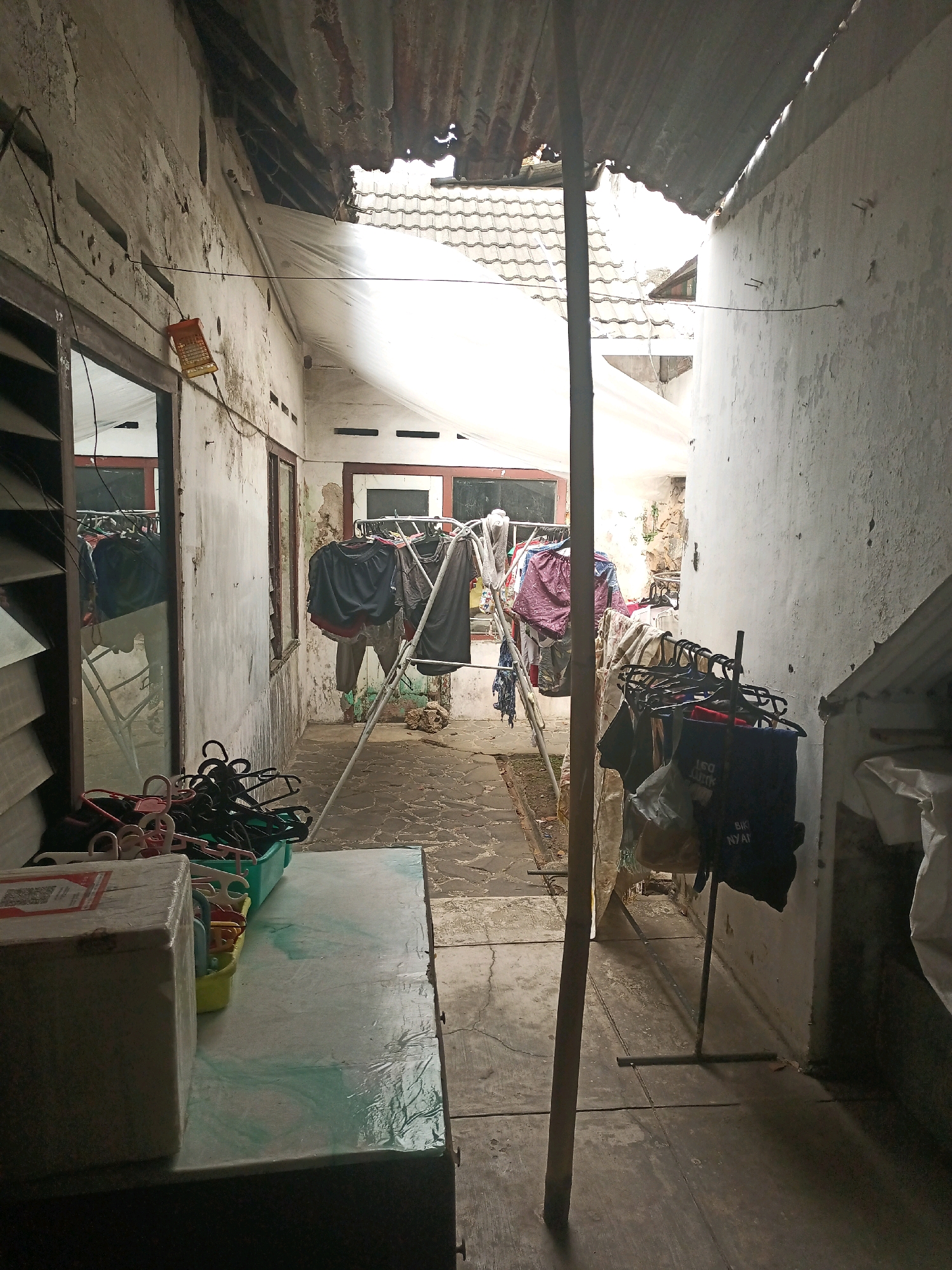 The image depicts a narrow, outdoor alleyway, likely a backyard or side passage, located in the city of Malang, Indonesia. The setting is characterized by worn, concrete flooring and textured, off-white walls that show signs of age and disrepair. A corrugated metal roof, partially damaged and exposing a white tarp underneath, covers part of the alley.
Several clotheslines are visible, laden with drying laundry, including dark-colored shirts, shorts, and patterned fabrics. Racks of hangers, some empty and others holding clothes, are also present. A green plastic container filled with hangers sits on a box-like structure in the foreground. A wooden pole stands vertically in the middle of the alley. Reflections of the drying clothes and the environment are visible in a large window to the left. The overall atmosphere is one of a functional, everyday space, likely a residential area given the presence of laundry. The lighting suggests it is daytime, with light filtering in from the open end of the alley. No people are visible in the scene.
