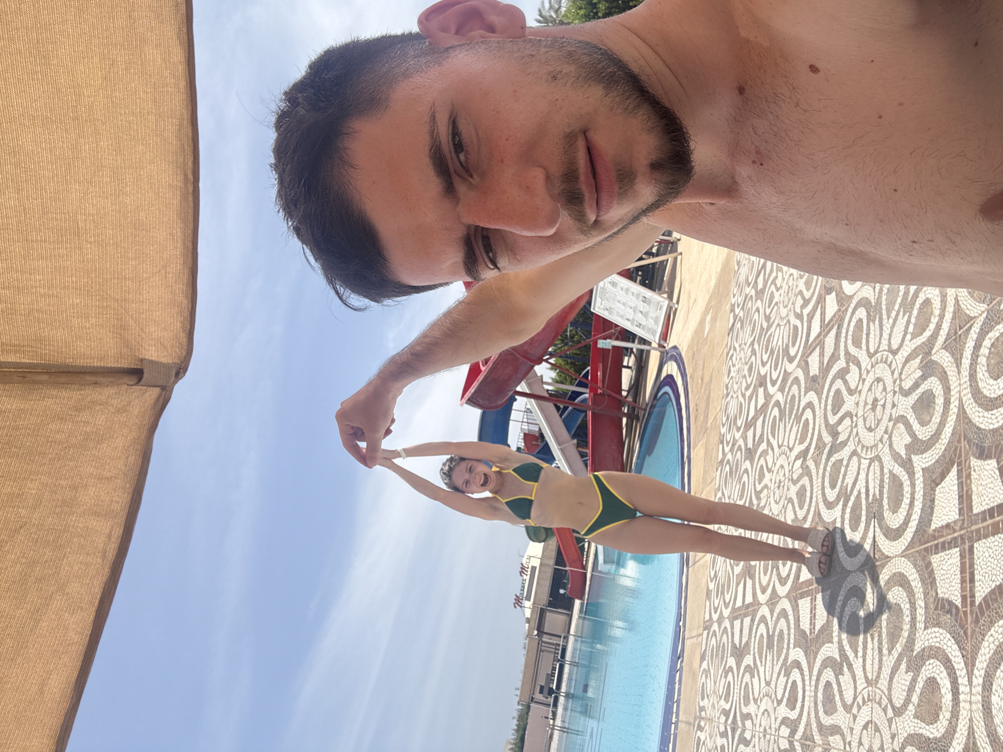 A man takes a selfie at a pool with a woman in a green and yellow swimsuit. The pool area features a waterslide and patterned tile flooring. A building labeled "Marina Mica" is visible in the background. The location is Unknown, Unknown.