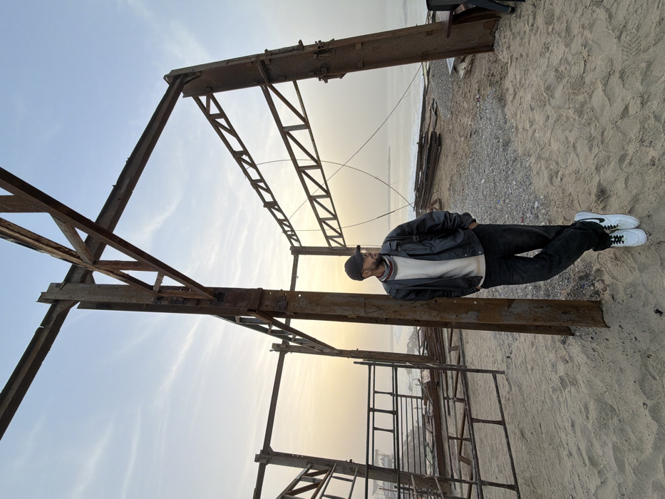 A man in a black jacket, tan sweater, dark pants, and white shoes stands on a sandy surface near rusty, derelict metal structures. The framework is extensive, reaching toward the sky with an open design. A stretch of water, possibly the ocean, is visible in the background under a soft, hazy sky. The scene conveys a sense of abandonment.