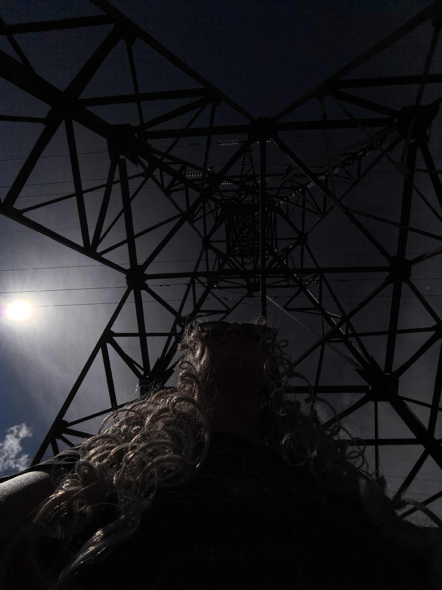 This is a low-angle, silhouette shot of a person standing beneath a large electrical pylon against a dusky sky. The pylon's intricate metal lattice work dominates the upper portion of the frame, creating a stark, geometric pattern that tapers towards the sky. Sunlight, visible as a bright orb on the left, casts a glare and illuminates some of the pylon's structure, as well as a few wisps of clouds.
The lower half of the image is darker, with the person's head and shoulders visible in silhouette. They have long, curly, light-colored hair that is caught by the light and some wind, giving it a dynamic, almost ethereal appearance. A clear plastic visor or safety glasses are perched on their head, adding a hint of detail to the obscured face. The overall mood is one of dramatic contrast and perhaps a sense of awe or insignificance in the face of the imposing industrial structure and the vastness of the sky. The time of day appears to be late afternoon or early evening, judging by the quality of the light and the dusky sky. No text is visible in the image.