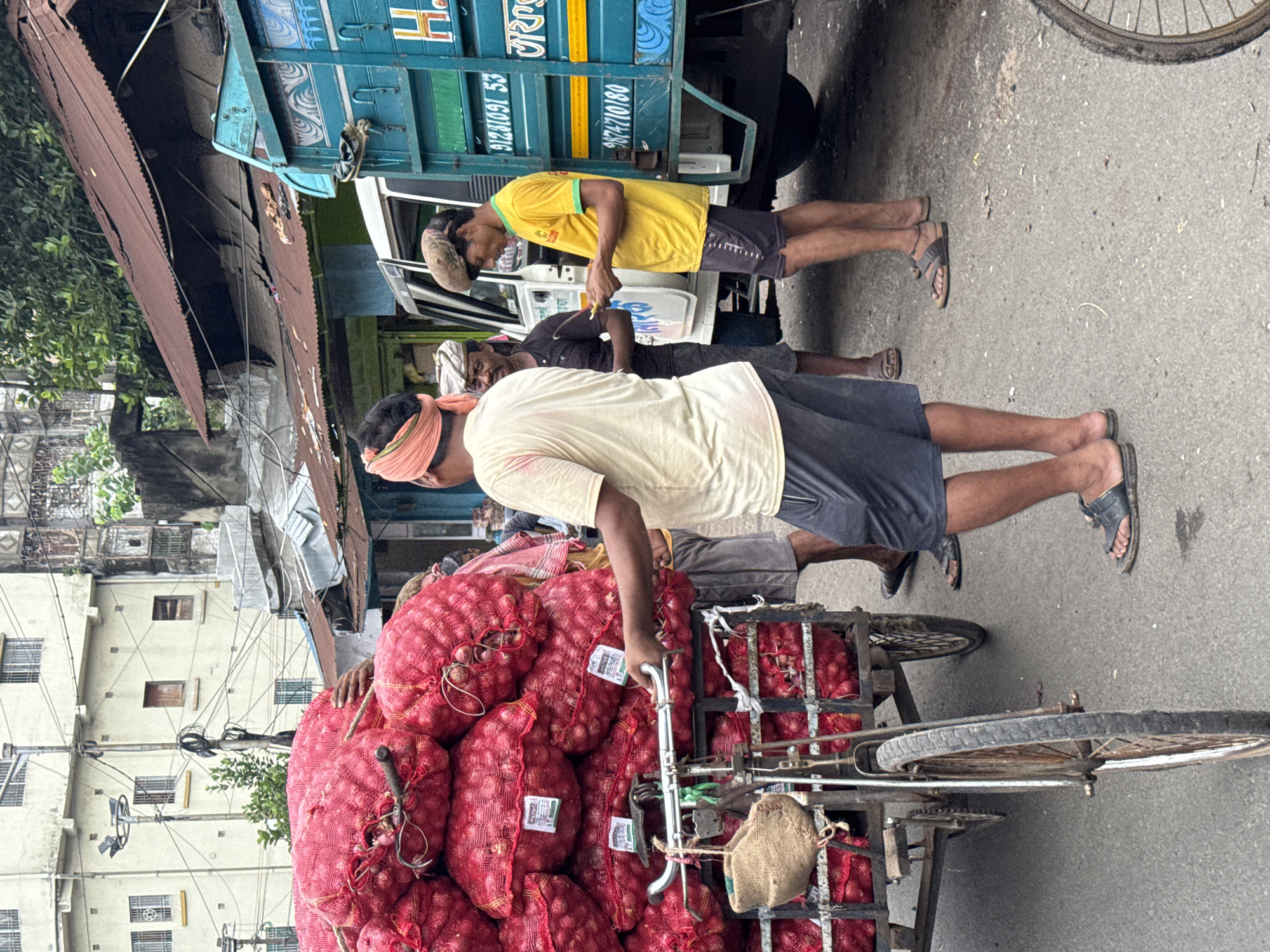 In a busy street scene in Kolkata, India, a man wearing a light yellow t-shirt and dark shorts is looking down at a mobile phone. He has a light-colored turban on his head and is standing next to a white truck with a teal-colored cargo section. The truck has writing on its side, including numbers and what appears to be Bengali script. In front of the truck, a cart piled high with large red net bags, presumably containing onions or similar produce, is visible. A bicycle with a woven bag hanging from its handlebars is attached to the front of this cart. Other people are present in the scene, including one individual partially visible in a turban and dark shirt, and another in a light orange headscarf and a light-colored t-shirt, holding onto the cart. The background shows buildings with multiple windows and overhead wires, typical of a bustling urban environment. The ground is paved and shows some signs of wear and tear.