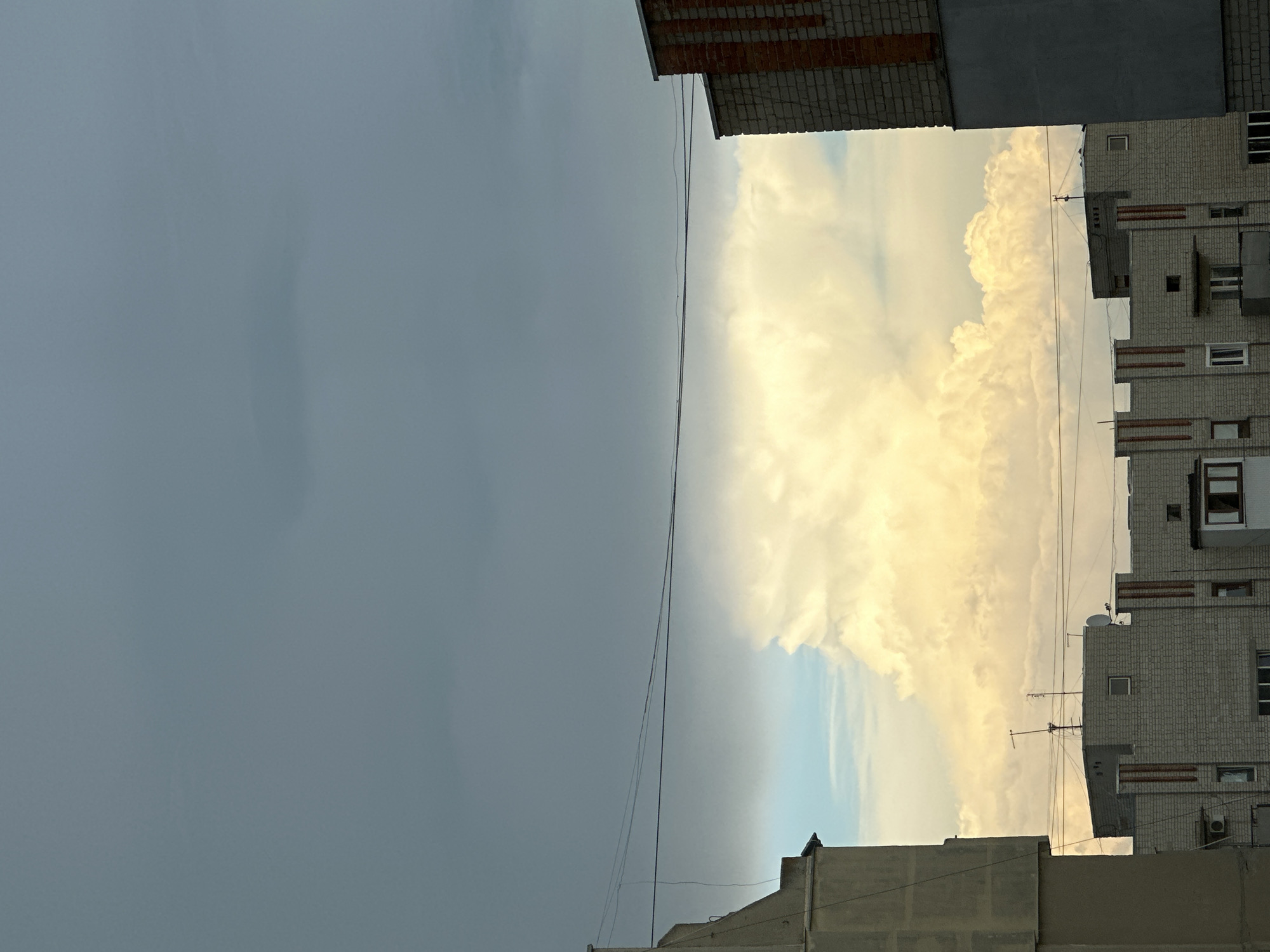 A view of a sky with large, bright cumulus clouds illuminated by sunlight, appearing to be at sunset or sunrise. The clouds are partially obscured by a dark, overcast sky above them. In the foreground, the tops of several apartment buildings are visible. One building on the left is a tall, grey concrete structure. On the right, a building made of red and grey brick is partially visible. Several apartment buildings with light grey brick exteriors and vertical red stripes are in the middle ground, some with visible antennas and satellite dishes on their roofs. Utility wires are strung across the scene. The overall impression is of an urban setting under a dramatic sky.