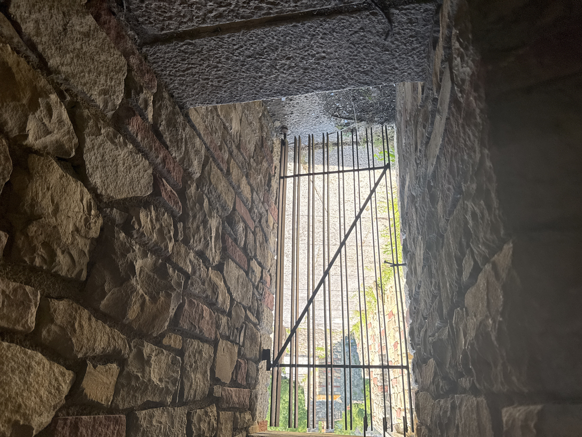 The image shows a stone stairway leading to a barred opening, suggesting the entrance to a dungeon or old cellar in the city of Veliko Tarnovo, Bulgaria. The walls are constructed of rough-hewn, multi-colored stones, typical of historical Bulgarian architecture. The stairway itself is also made of large, rough-cut stone blocks. Beyond the bars, a path is visible, lined with similar stone walls, leading into a brighter area with greenery. The scene evokes a sense of history and potentially confinement.