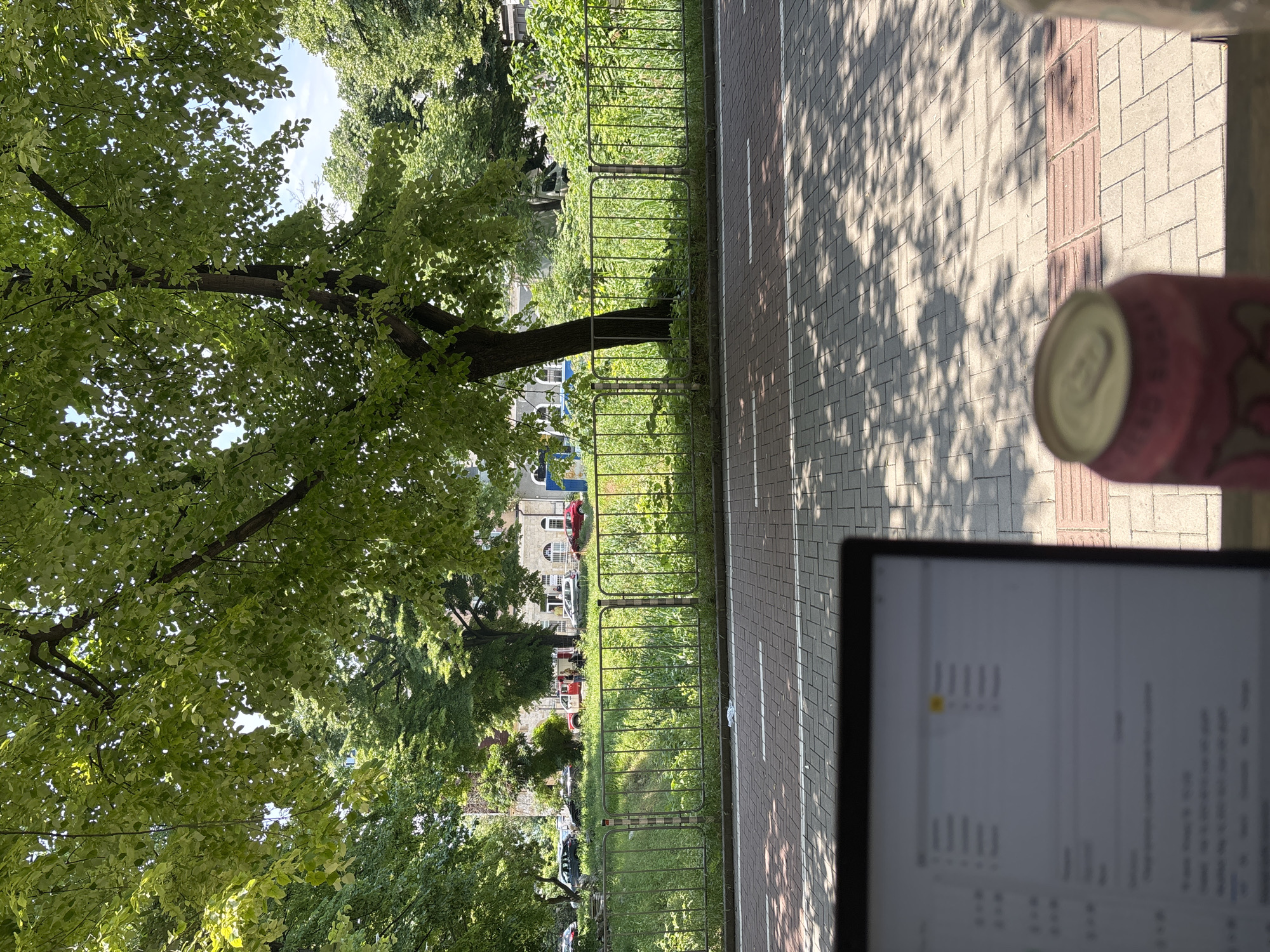 The image, if rotated 90 degrees clockwise, depicts an outdoor scene in Varna, Bulgaria, characterized by a mix of natural greenery and urban infrastructure. In the immediate foreground, a blurry pink beverage can rests beside a partially visible laptop screen. The screen displays what appears to be lines of text or code, too indistinct to decipher.
A paved pathway, constructed from light-colored rectangular pavers, stretches across the lower portion of the scene. It features a narrower strip of reddish-brown tactile pavers and a white dashed line. Distinct, dappled shadows from overhead trees fall across this path, indicating bright sunlight. A low metal fence separates this pathway from a lush, green area.
Beyond the fence, a dense canopy of vibrant green trees dominates the upper half of the image, with patches of blue sky visible through the foliage. In the distance, glimpses of multi-story buildings with architectural details, including arched windows, can be seen nestled among the trees. Several cars, including a prominent red vehicle, are parked or moving in the background near these structures.
The strong, well-defined shadows suggest a clear, sunny day, likely during the morning or afternoon. The overall impression is one of a peaceful, pleasant urban park or residential area, offering a tranquil setting for outdoor work or leisure.