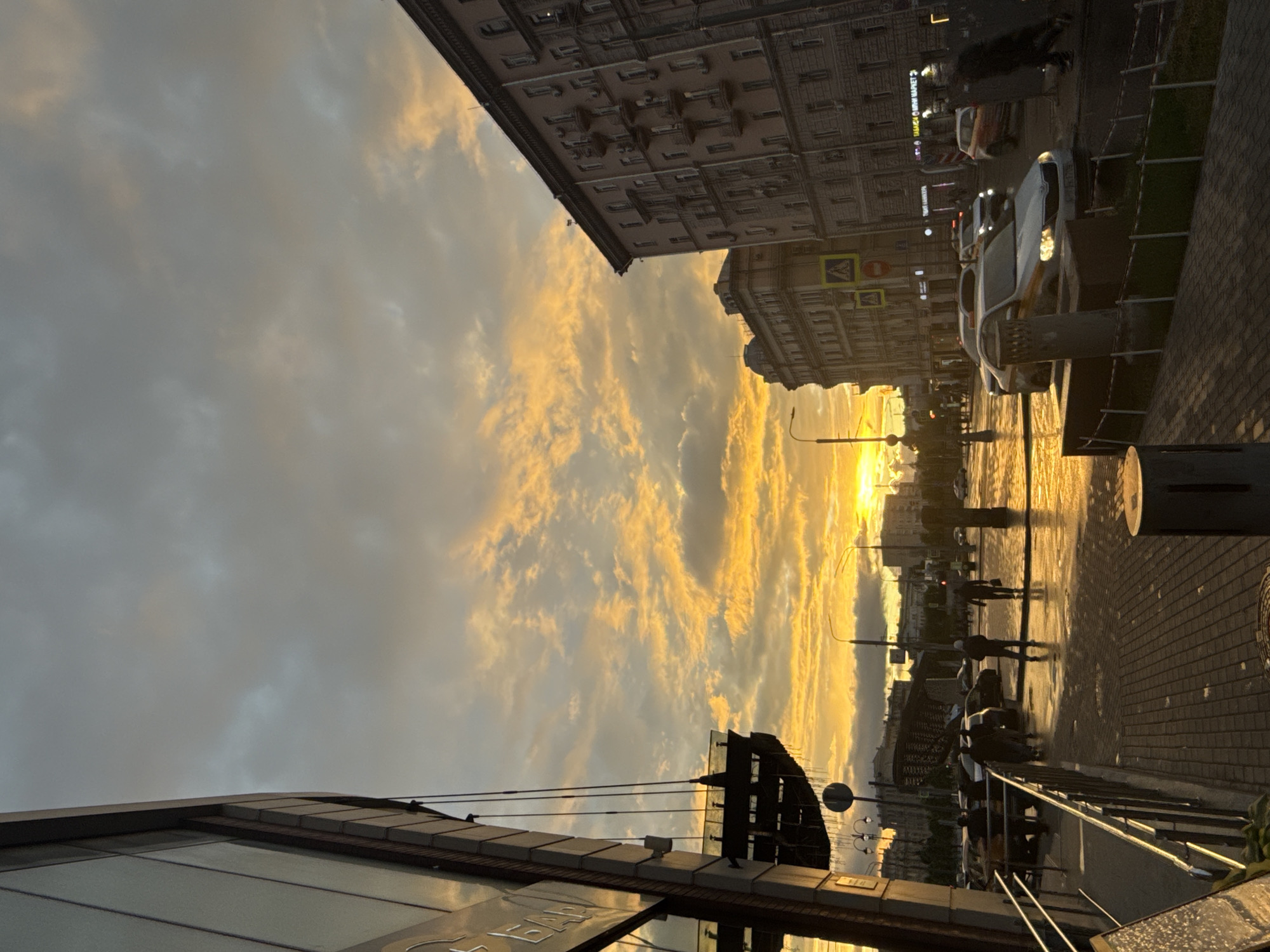 Sunset scene in Moscow, Russia. A street view captures buildings with multiple floors and windows, while traffic signals and street lamps stand along the road. Cars are parked alongside the road. Pedestrians walk along the sidewalk. The sky is filled with clouds illuminated by a vibrant sunset.