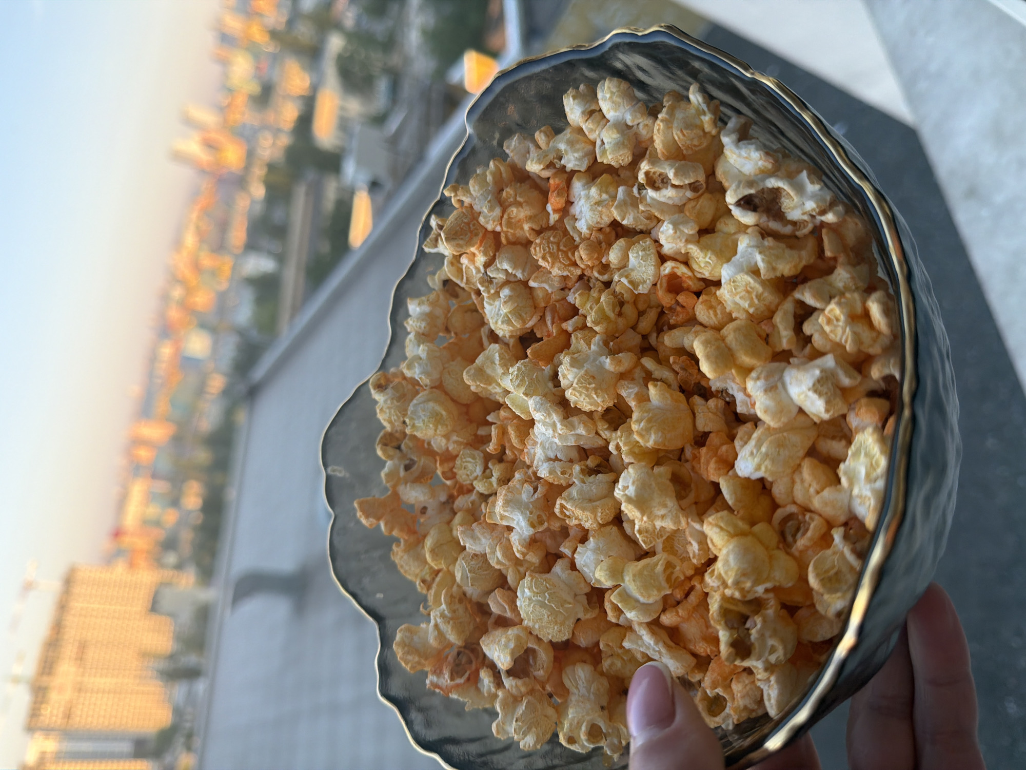 A hand holds a bowl of popcorn near a window overlooking a city. The bowl is dark gray with a gold rim and is filled with fluffy popcorn, some of which has an orange hue, possibly from seasoning. In the background, the cityscape of Kyiv, Ukraine, is visible at sunset. Buildings, including a tall construction crane, are silhouetted against the orange and blue sky. The scene suggests a casual moment of enjoying a snack while taking in the view of the city.