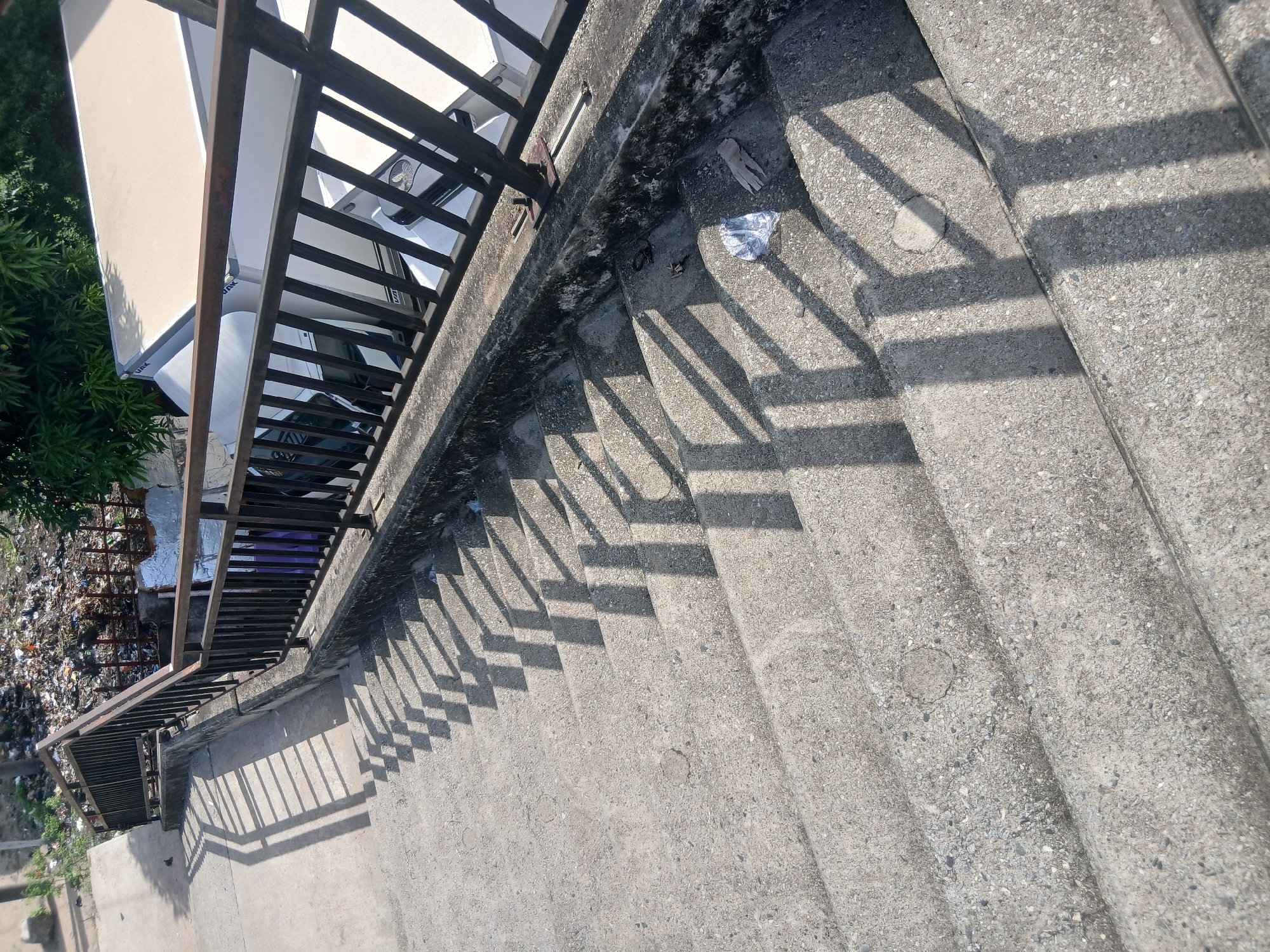 The image shows a concrete staircase with a dark metal railing on the left side. The stairs are exposed to direct sunlight, casting sharp shadows of the railing across the steps. A small pile of trash is visible to the left of the railing, including plastic bottles and general debris. Litter, consisting of gloves and plastic bags is strewn on the stairs. The location is strongly suspected to be Lagos, Nigeria due to commonalities in materials and environmental conditions consistent with documented locales in Lagos.