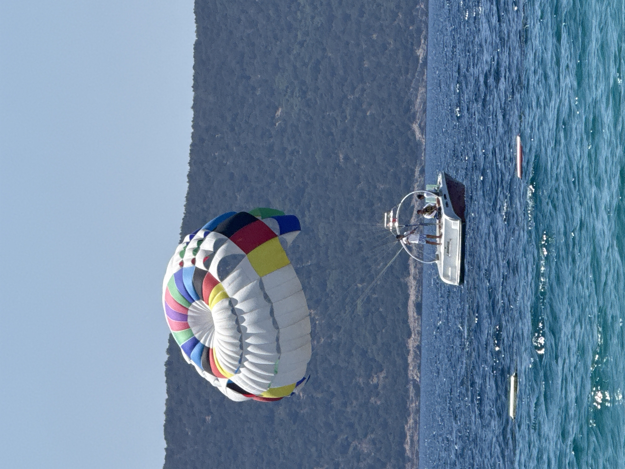 A parasail, with segments of red, yellow, blue, green, and purple, is aloft over a body of bright blue water. Below the parasail, a white boat with a dark red stripe along its waterline is visible. Three individuals are on the boat. One person stands at the back of the boat, while two others are seated. The boat is in motion, and it appears to be towing the parasail. In the background, a lush, dark green hillside rises from the water. The sky is a clear, pale blue. The location is the Aegean Sea, near Bodrum, Turkey.