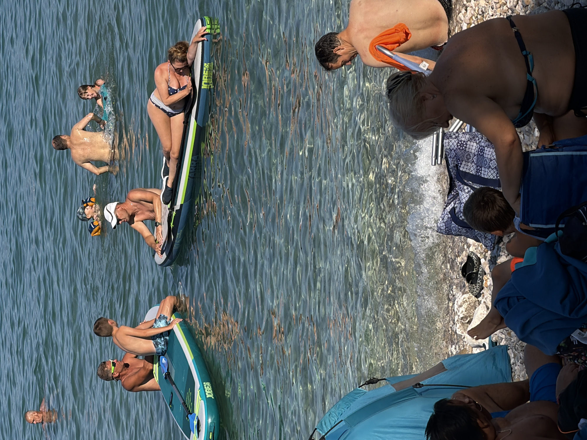 In Općina Baška, Croatia, a group of people are enjoying a day at the beach. The image captures a sunny scene with clear, shallow water. In the foreground, several individuals are seated on a pebble beach. One woman is wearing a dark bikini top and bottoms, with her hair tied back. Beside her, a person wearing blue swim trunks is looking down. To the left, a large, blue tent-like structure is partially visible.
Further out in the water, a woman in a striped bikini lies on a paddleboard. Another person, also on a paddleboard, is seated and looking towards the right. A man with sunglasses is on a paddleboard with a boy. In the background, other individuals are swimming and playing in the water. One person is wearing an orange life vest. The water is transparent, revealing the pebbly seabed.
