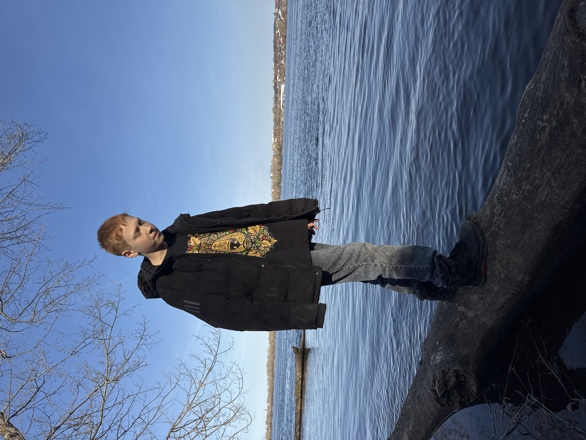 A fair-skinned person with reddish hair stands on a fallen tree trunk overhanging a body of water under a bright blue sky. The person wears a black puffer jacket over a black T-shirt with a gold design on the chest, blue jeans, and black shoes with red detailing. The water reflects the sky, and a distant shoreline is visible. Bare tree branches are visible in the lower left corner. The location is implied to be in the city of Noviy, Russia, based on the prompt.