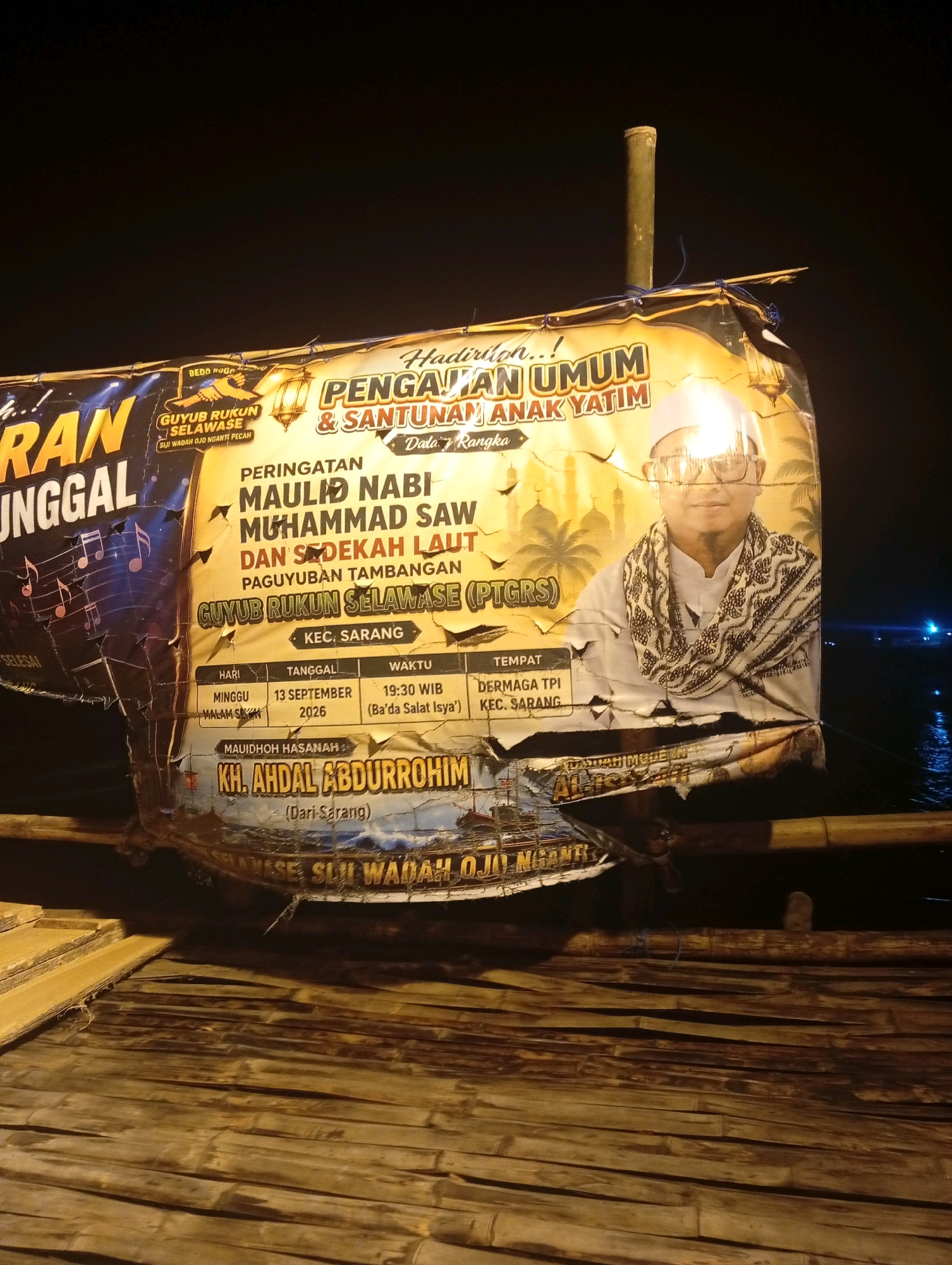 This image depicts a nighttime scene in the city of Banowan, Indonesia, featuring a large, damaged banner displayed outdoors. The banner, which appears to be made of plastic or vinyl, is illuminated by an unseen light source, highlighting its text and imagery. The banner is attached to a wooden structure and is partially torn and weathered, indicating exposure to the elements.
The banner's content includes text in Indonesian, advertising a public recitation and donation event for orphans. Key phrases visible are "PENGAJIAN UMUM & SANTUNAN ANAK YATIM" (Public Recitation & Orphan Donation) and "PERINGATAN MAULID NABI MUHAMMAD SAW" (Commemoration of the Prophet Muhammad's Birthday). There is also information about the date and time of the event: "MINGGU 13 SEPTEMBER 2026" (Sunday, September 13, 2026) and "19:30 WIB" (7:30 PM Western Indonesian Time). The location is stated as "DERMAGA TPI KEC. SARANG" (TPI Pier, Sarang District). A photograph of a man wearing a white cap and robe is featured prominently on the banner, along with decorative elements like a lantern and minarets. Below the main event details, the name "KH. AHDAL ABDURROHIM" is listed as the speaker or honoree, with "(Dari Sarang)" indicating his origin.
The surrounding environment is dark, suggesting it is night. The banner is suspended over a wooden platform or walkway made of bamboo, which is also illuminated by the same light source. In the background, a body of water is visible, with some distant lights reflecting on its surface. The overall impression is of a community event being advertised in an outdoor, possibly seaside or riverside, location.