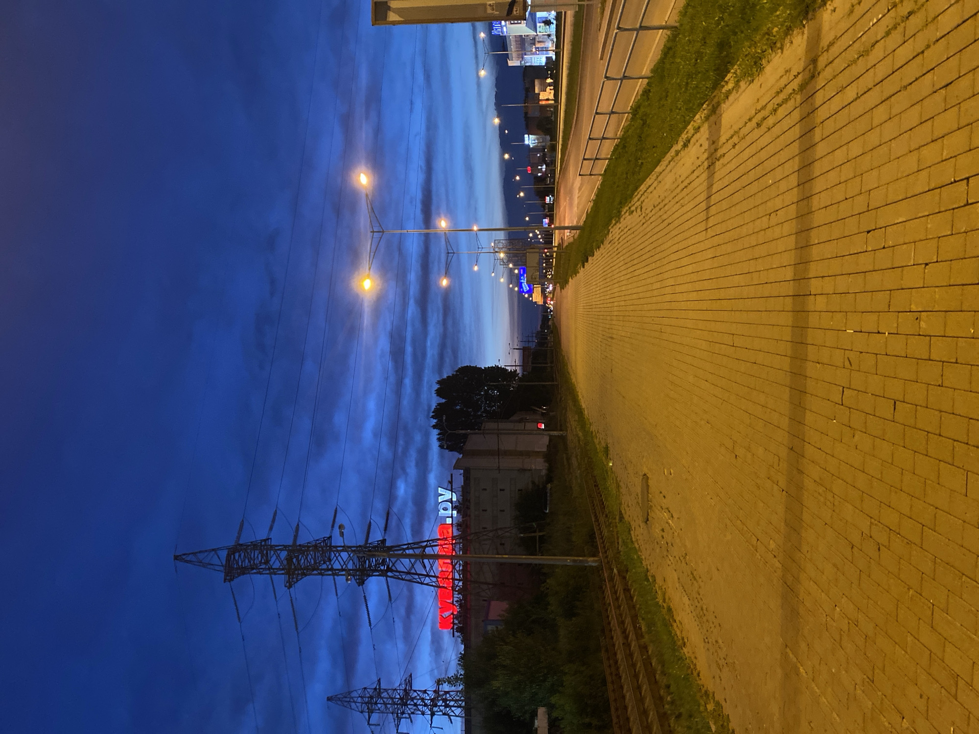 The image depicts an urban scene at dusk or dawn in Samara, Russia. A wide, yellow brick sidewalk runs parallel to a set of train tracks on the left and a road with streetlights on the right. In the background, a large building is illuminated by red neon signs that read "Куйбышев.py" and some lighter blue lettering. High-voltage power line towers are visible against the deep blue, cloudy sky. The road is lined with streetlights, and distant buildings with illuminated signs are visible. There are no people or vehicles clearly identifiable in the image. The environment appears to be an industrial or commercial area on the outskirts of the city.