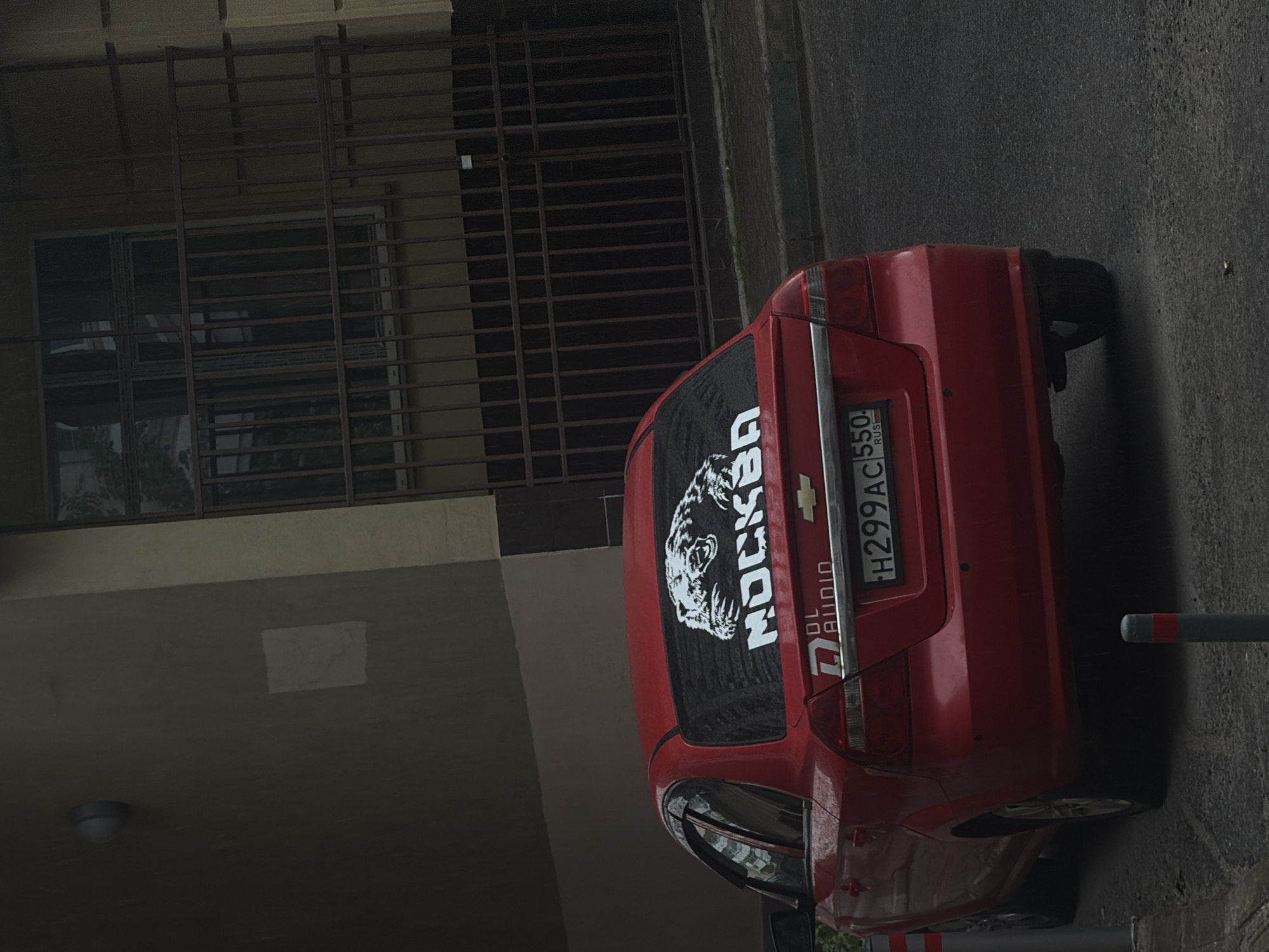 A red Chevrolet Aveo is parked on a street in Moscow, Russia. The car has a sticker on its rear window that says "MOSKVA" with a picture of a bear. The license plate reads "H 299 AC 550. RUS". The car is parked next to a building with a barred window and a light fixture on the wall. The street is asphalt, and there are two bollards in the foreground. The scene appears to be overcast with visible raindrops on the car and the ground.