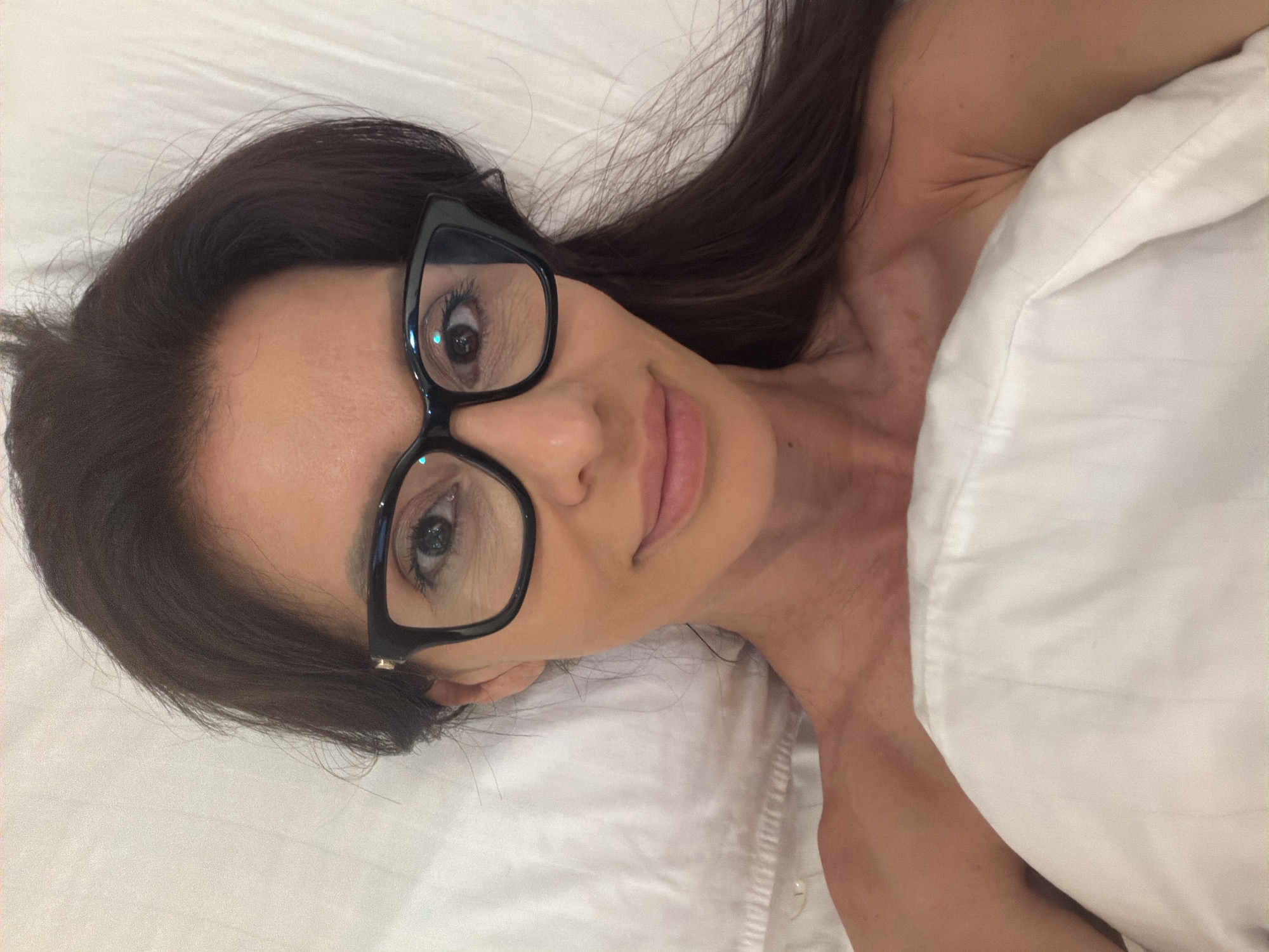 A woman with dark hair and glasses lies in a bed, her upper body covered by a white comforter. The room appears to be a bedroom with white linens. There is no indication of the city of Varna, Bulgaria.