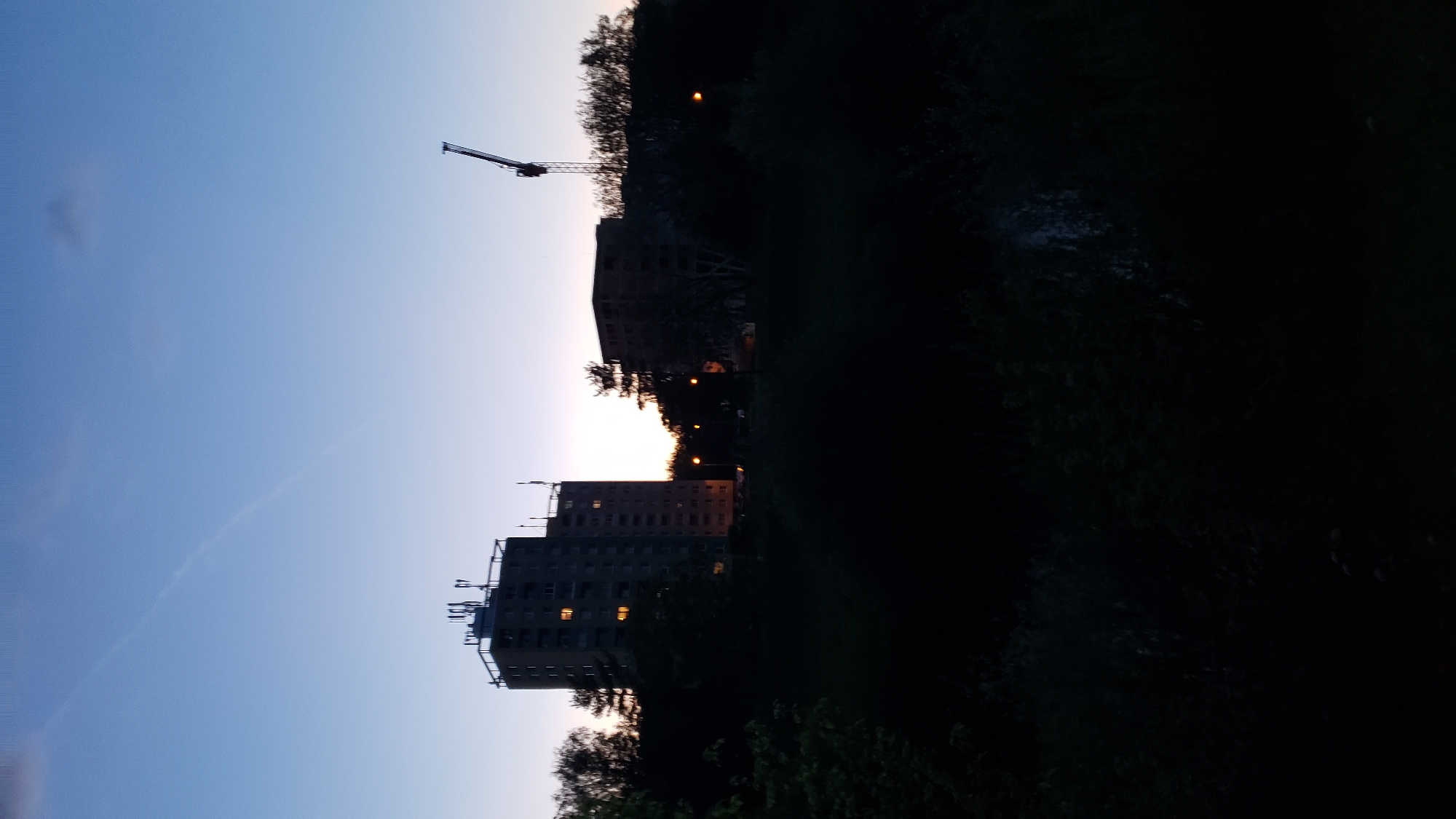 At dusk, two silhouetted apartment buildings stand in the distance, their windows illuminated by interior lights. To the right of these buildings, a construction crane is visible against the fading sky. A gradient of blue and purple colors the sky, with a faint contrail streaking across the upper portion. In the foreground, a dark expanse of trees and foliage obscures the ground. This scene is located in the city of Prešov, Slovakia.