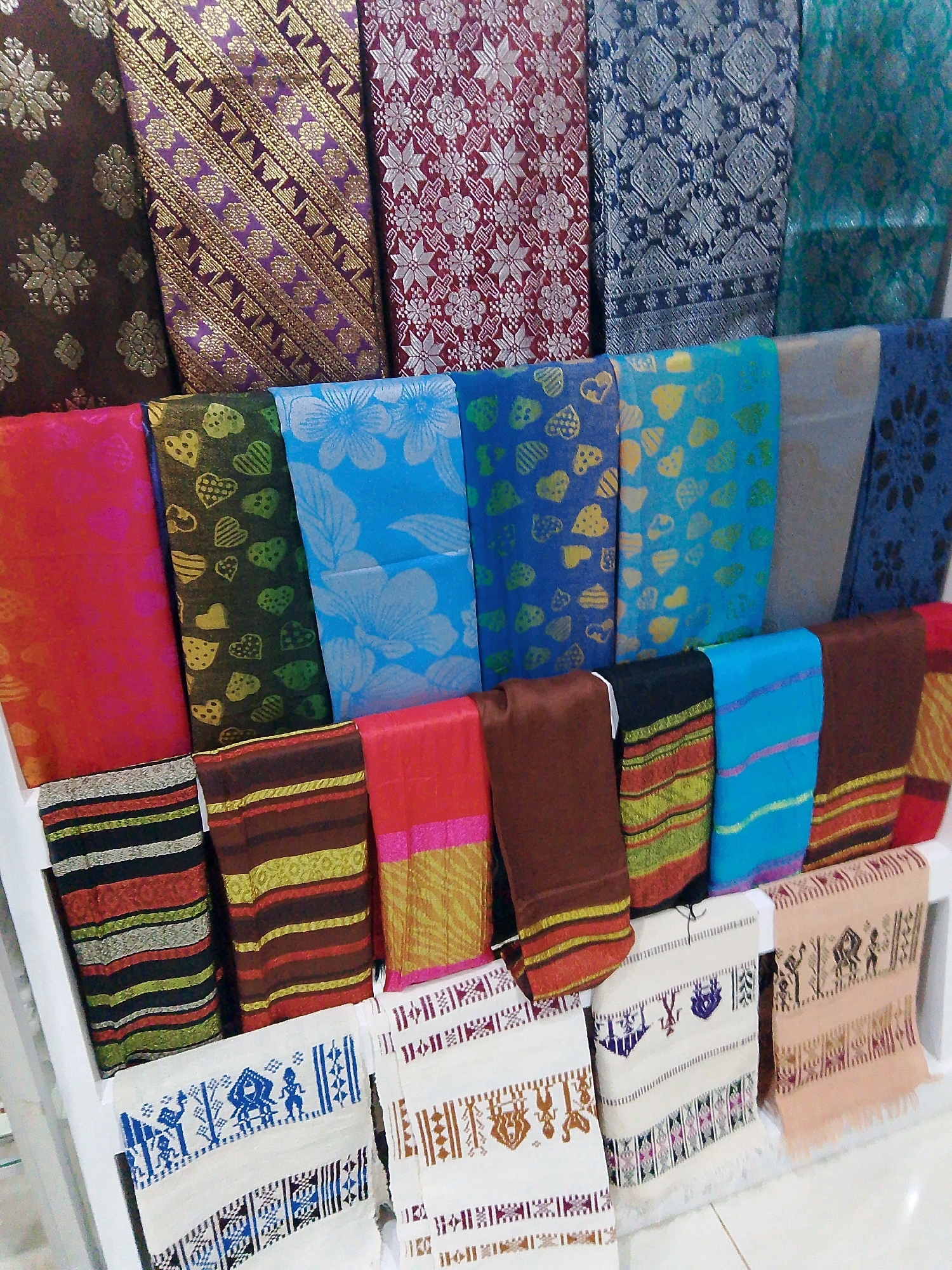 The image displays a retail setting in Bunkate, Indonesia, showcasing a vibrant collection of Indonesian textiles, likely sarongs or fabrics. The textiles are arranged on a tiered display rack, with a variety of patterns and colors. The top row features richly patterned fabrics in deep reds, purples, blues, and greens, with intricate woven designs. Below, the second row consists of brightly colored fabrics, including fuchsia, lime green with heart patterns, and various shades of blue with floral motifs. The bottom row presents textiles with striped patterns in earthy tones, along with white fabrics featuring embroidered designs and fringed edges. The scene appears to be an indoor market or shop, with the textiles illuminated by artificial lighting. No people are visible in the image.