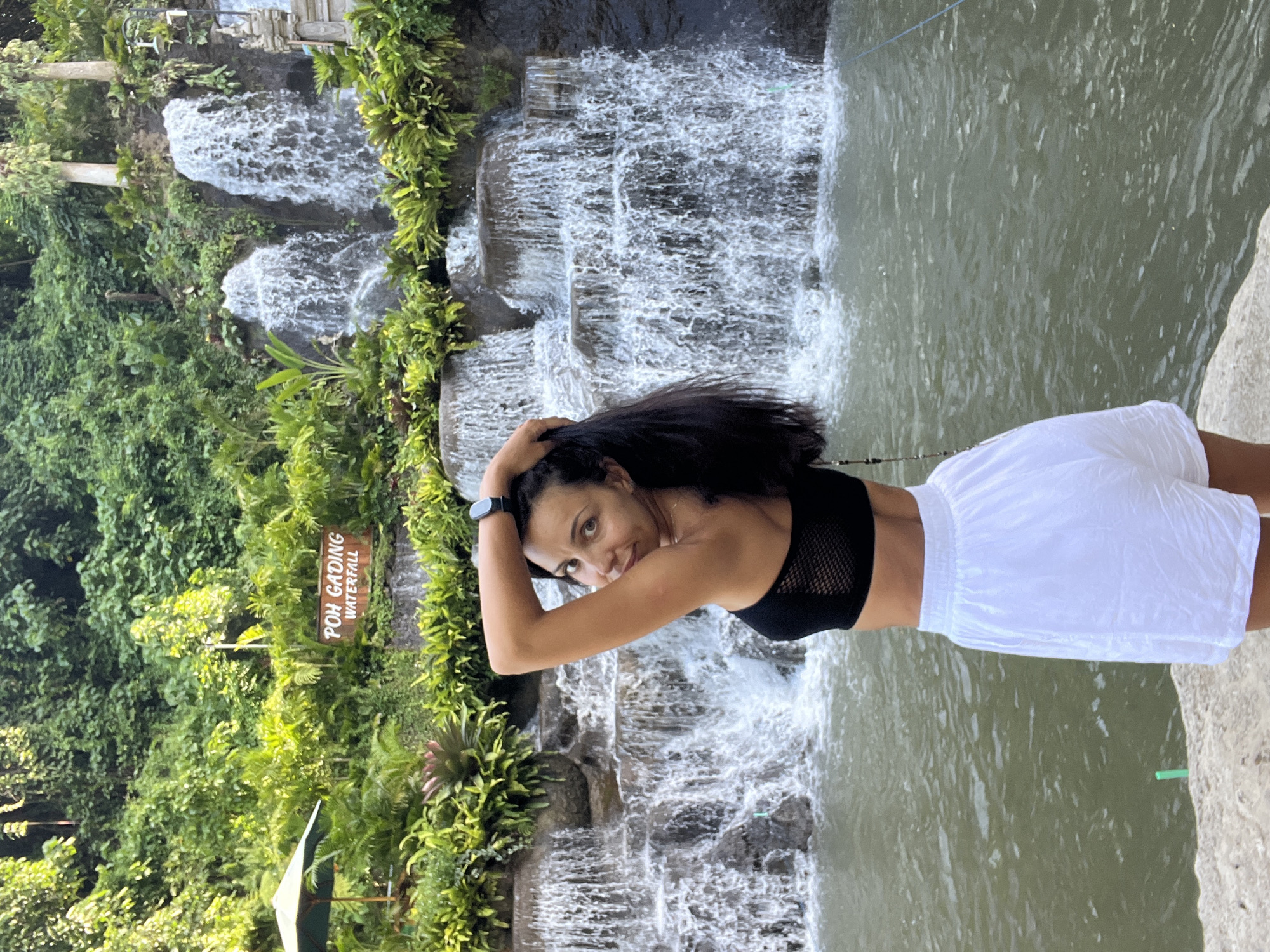 The image shows a woman standing in front of a waterfall. She is wearing a black top and white shorts and has dark hair. She has her hands on her head and is looking at the camera. The waterfall is multi-tiered with water flowing down the rocks, which are covered in green plants. A sign that reads "Poh Gading Waterfall" is visible on the left side of the frame. The body of water at the top of the waterfall is murky. The location is Sukawati, Indonesia.