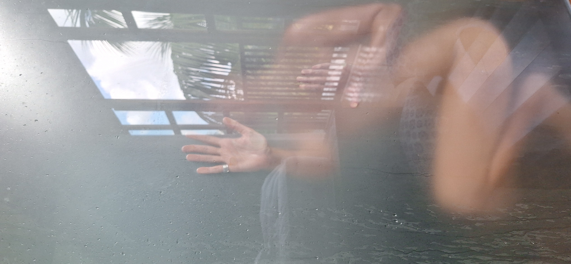 The image captures a person's hand pressed against a wet, frosted glass surface, possibly a shower door, with water droplets visible. Through the glass, a blurry reflection shows a portion of the person's body and surroundings. The background outside the glass appears to be a balcony or outdoor area with railings and palm fronds, suggesting a tropical environment. The lighting indicates it might be daytime, with sunlight filtering through the windows and palm leaves. The scene suggests an intimate or private moment, possibly in a residence or hotel. The location in Payangan, Indonesia, implies a tropical setting with lush vegetation.