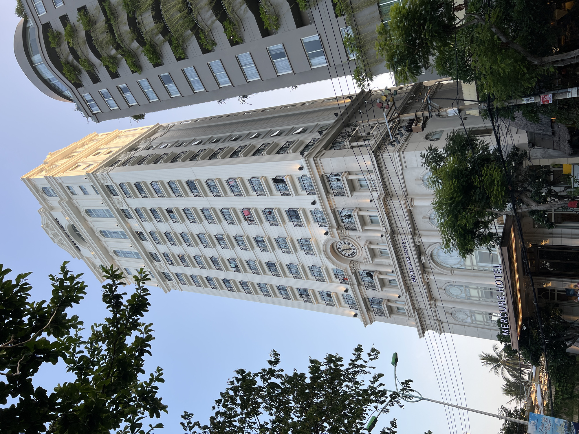 The image depicts the Mercure Hotel in Nha Trang, Vietnam, a tall, white, ornate building with a clock face and balconies. The hotel is situated next to a modern gray building with green, tiered balconies. Trees and overhead power lines are visible in the foreground. The sky is clear and blue. A street lamp is also present in the foreground. The hotel's name, "MERCURE HOTEL," is illuminated in prominent signage at the entrance.