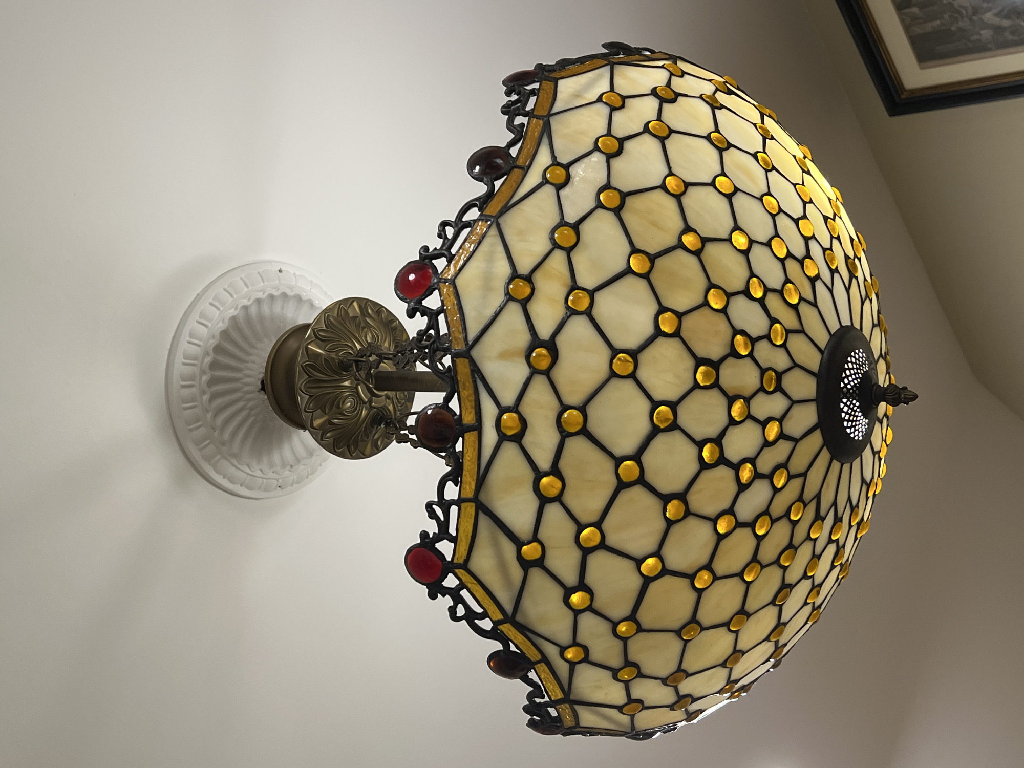The image shows a close-up, upward view of a Tiffany-style stained glass ceiling light fixture. The light is illuminated and casts a warm glow through the cream-colored glass panels, which are arranged in a hexagonal pattern outlined by dark solder lines. Small, round yellow glass beads are set at the intersections of the solder lines. The decorative metal frame surrounding the stained glass is dark, with ornate scrollwork along the top edge, adorned with several red and dark red glass cabochons. The fixture is suspended from a white, ornate ceiling medallion. A dark metal finial hangs from the bottom of the fixture, with a perforated design. The background is a plain white ceiling. The location provided is Kallithea, Greece, but there are no visual cues in the image to confirm this specific location; the scene appears to be an interior setting. There are no people or actions depicted.