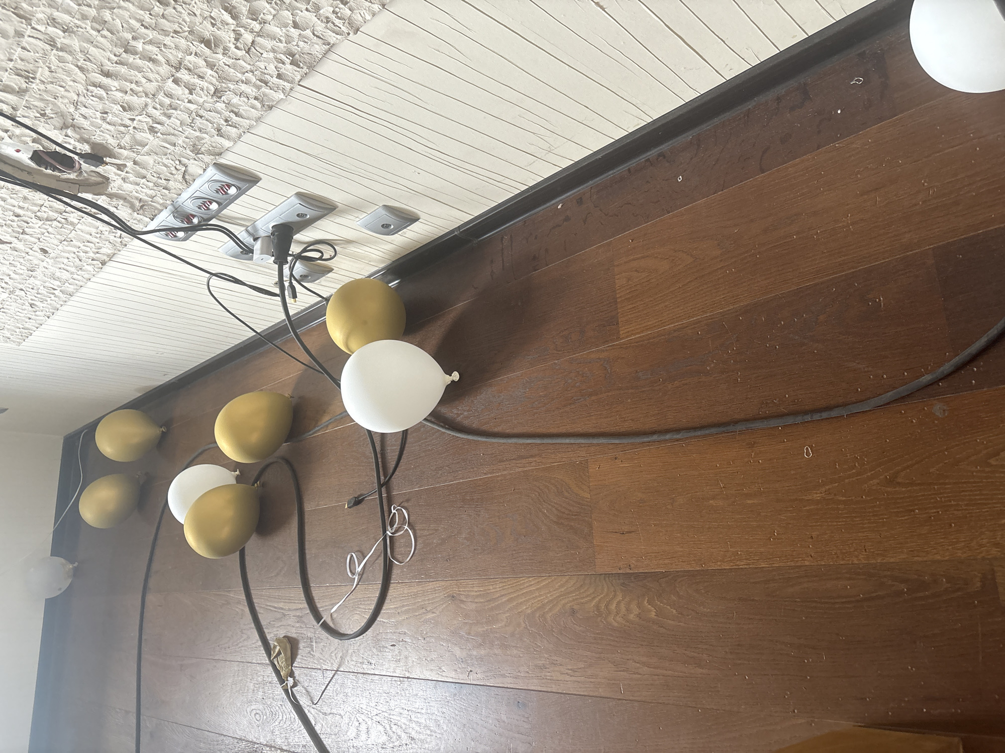 The image depicts a dimly lit interior space with a dark wooden floor. A series of gold and white balloons are scattered across the floor, some deflated and others still inflated. Black electrical cords snake across the floor and up a textured, off-white wall. The wall features multiple electrical outlets and wiring, suggesting an unfinished or in-progress renovation. The overall impression is one of disarray and perhaps a party that has recently concluded or is being set up. The location is identified as Orekhovka, Russia, but no specific landmarks or city features are visible to confirm this.