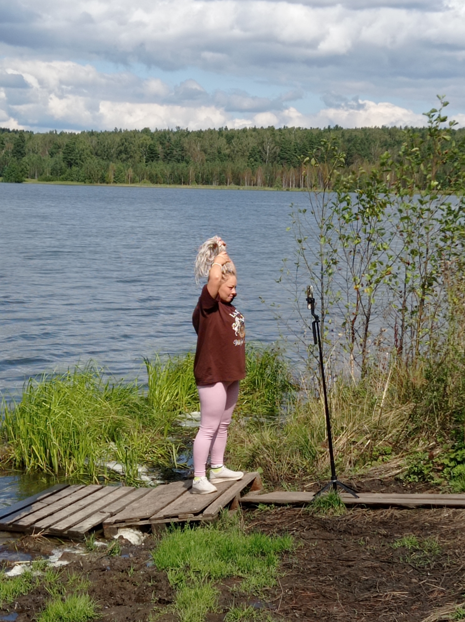 A person stands on a wooden platform by a body of water. The person has blonde hair pulled up in a bun and is wearing a brown t-shirt and pink leggings. They are holding their hands up near their head. Behind them, a forest lines the far shore of the water. The sky is cloudy. The location appears to be the shore of Lake Sukhoy Log in Degtyarsk, Russia.