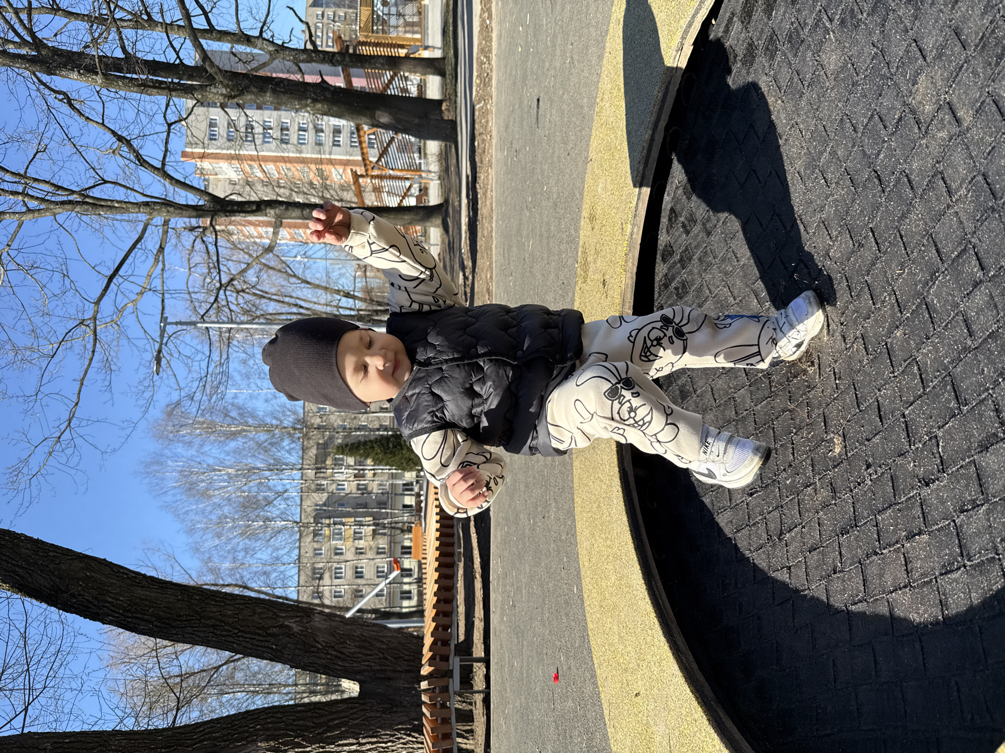 A young child lies on their back in a playground in Rusanovo, Russia. The child wears a dark beanie, a black puffer vest, and a patterned long-sleeved shirt and pants, paired with white sneakers. They are positioned on a round, partially shaded surface that features a black textured area and a surrounding yellow border. The playground is bordered by bare trees, and in the background, buildings can be seen. The weather appears sunny, with a clear blue sky visible through the tree branches.