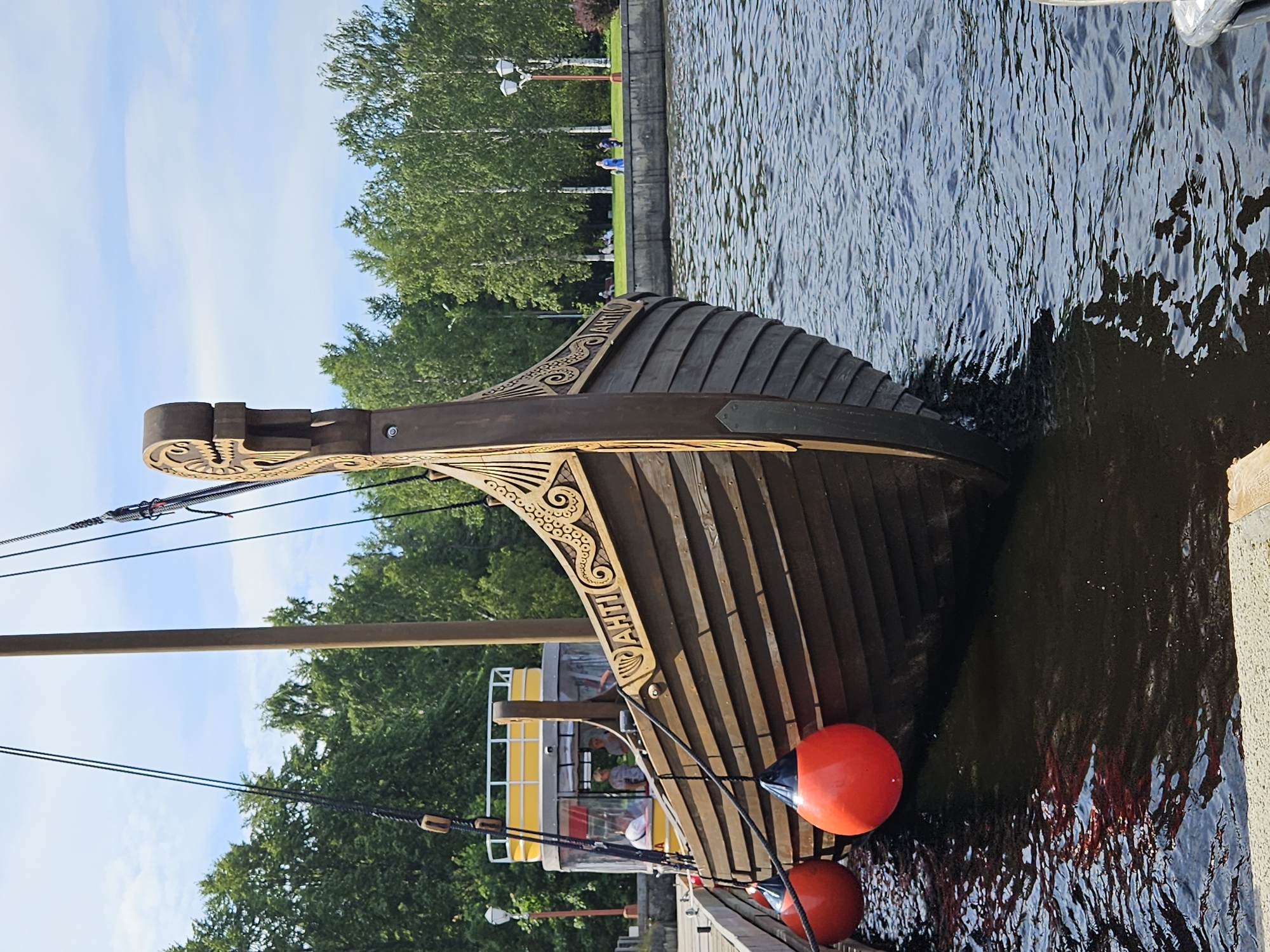 A wooden Viking-style ship is docked at a waterfront with a yellow and white boat in the background. The ship has intricate carvings along its side and a dragon-head figure at its bow. Two large orange buoys hang from the side of the ship. Trees line the waterfront in the background, and a few people are visible in the distance, some sitting on the grass and others walking. The sky is partly cloudy. The scene appears to be a tourist attraction or recreational area.