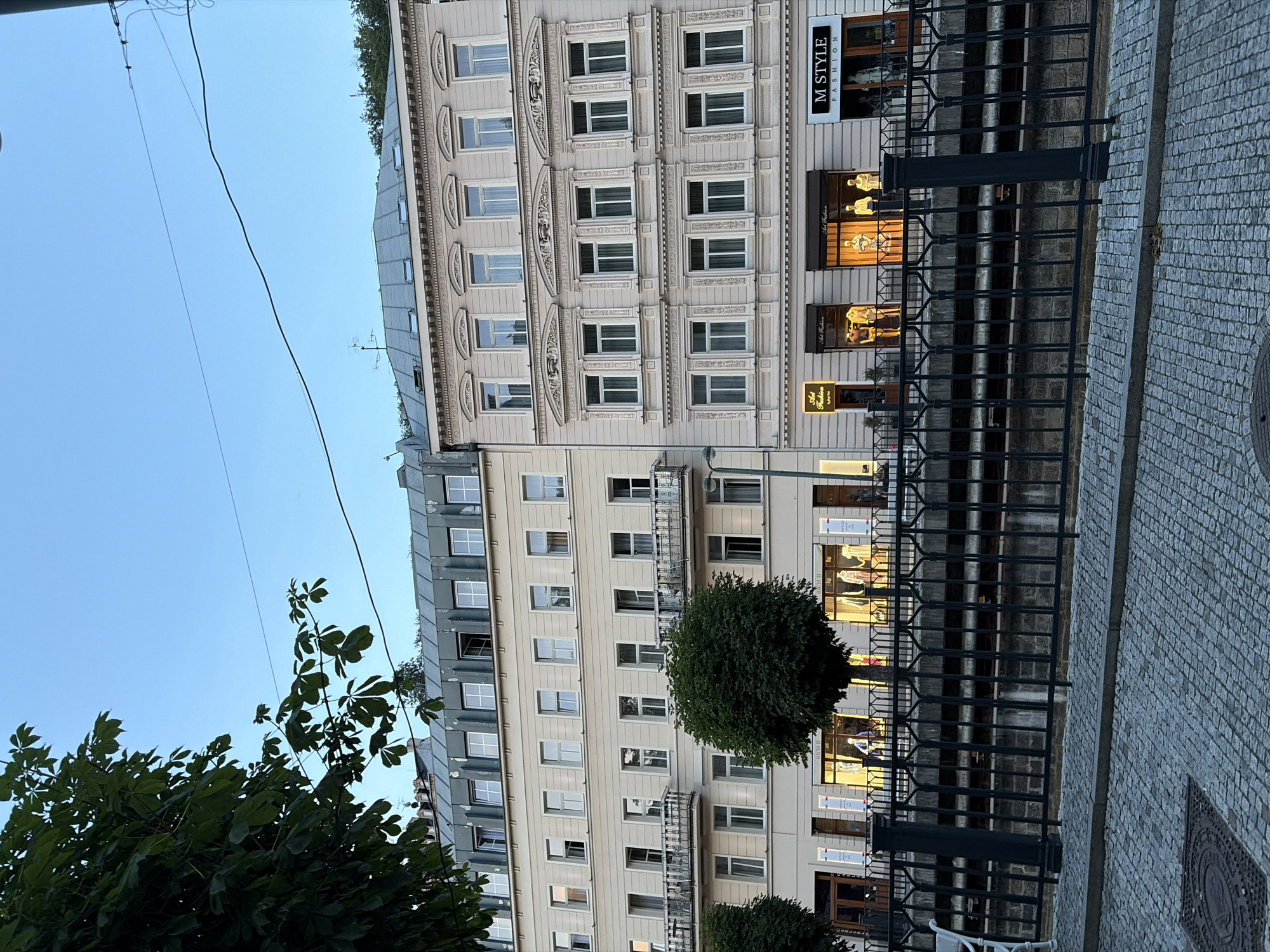 The image shows a street view in Karlovy Vary, Czechia. In the foreground, a cobblestone street and a black metal fence with ornate designs are visible. Beyond the fence, a body of water flows, lined with stone walls. Two multi-story buildings with light-colored facades dominate the background. The building on the left has a grey, mansard-style roof with dormer windows and several balconies on its upper floors. The building on the right features a more decorative facade with elaborate window frames and a corniced roofline. The ground floors of both buildings appear to house retail establishments, with illuminated shop windows displaying mannequins and merchandise. Signs such as "Art Fashion" and "M STYLE FASHION" are visible above the shops. Trees with green foliage are present in front of the buildings and to the left of the frame. The sky is a clear, light blue, suggesting it is either early morning or late evening. There are no people clearly discernible in the image, only mannequins in the shop windows. The overall scene depicts a European city street with commercial activity and architectural heritage.