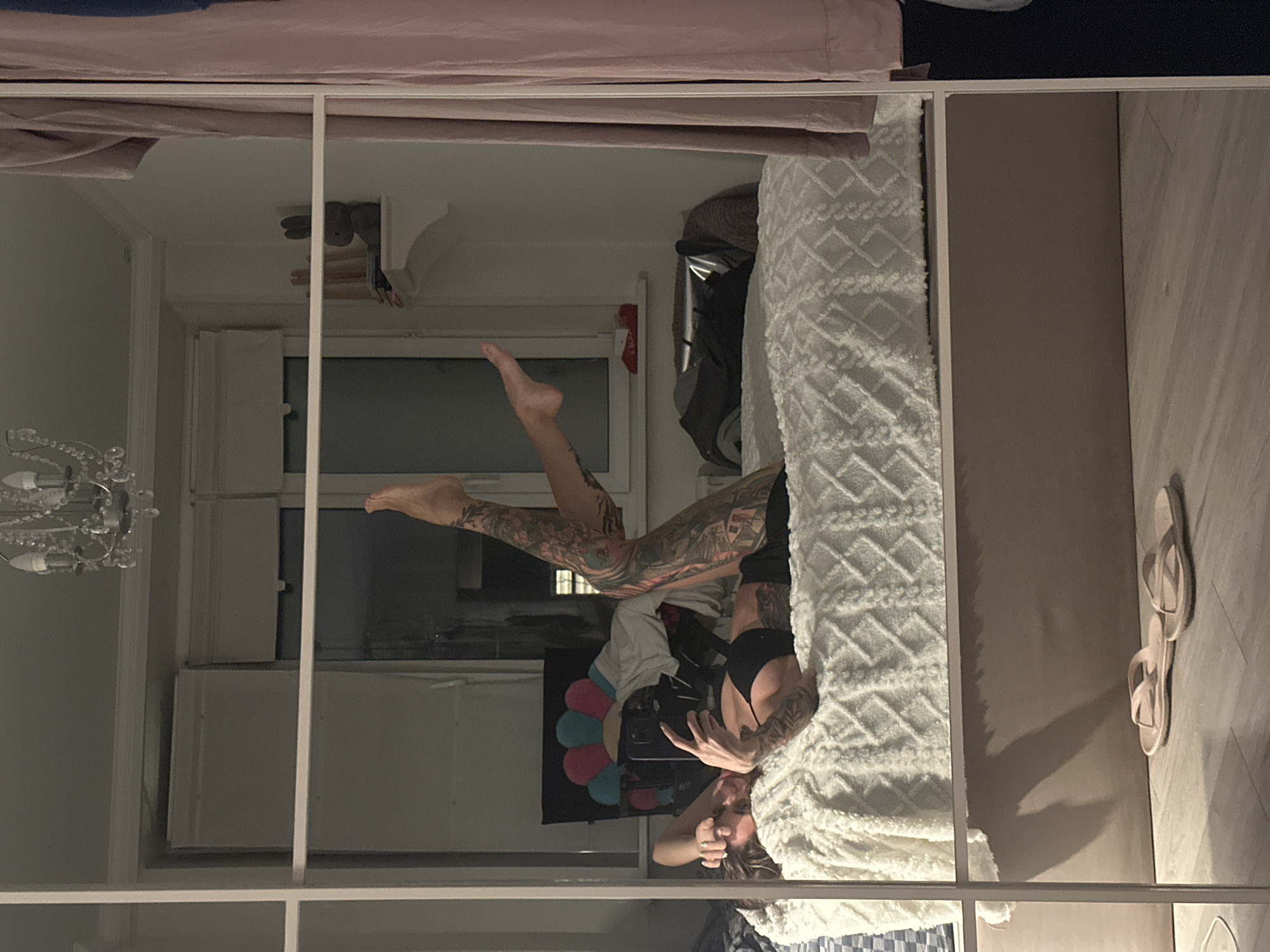 The image shows a woman lying on a bed in what appears to be a bedroom. She is taking a selfie with her phone. She has tattoos on her arms and legs. The bed is covered with a white blanket. There are clothes on a rack to the right of the bed. There is a mirror in the foreground, reflecting the scene. The overall impression is that this is a candid, personal moment captured in a private space, likely an apartment. The location in Moscow, Russia is not discernible from the image.