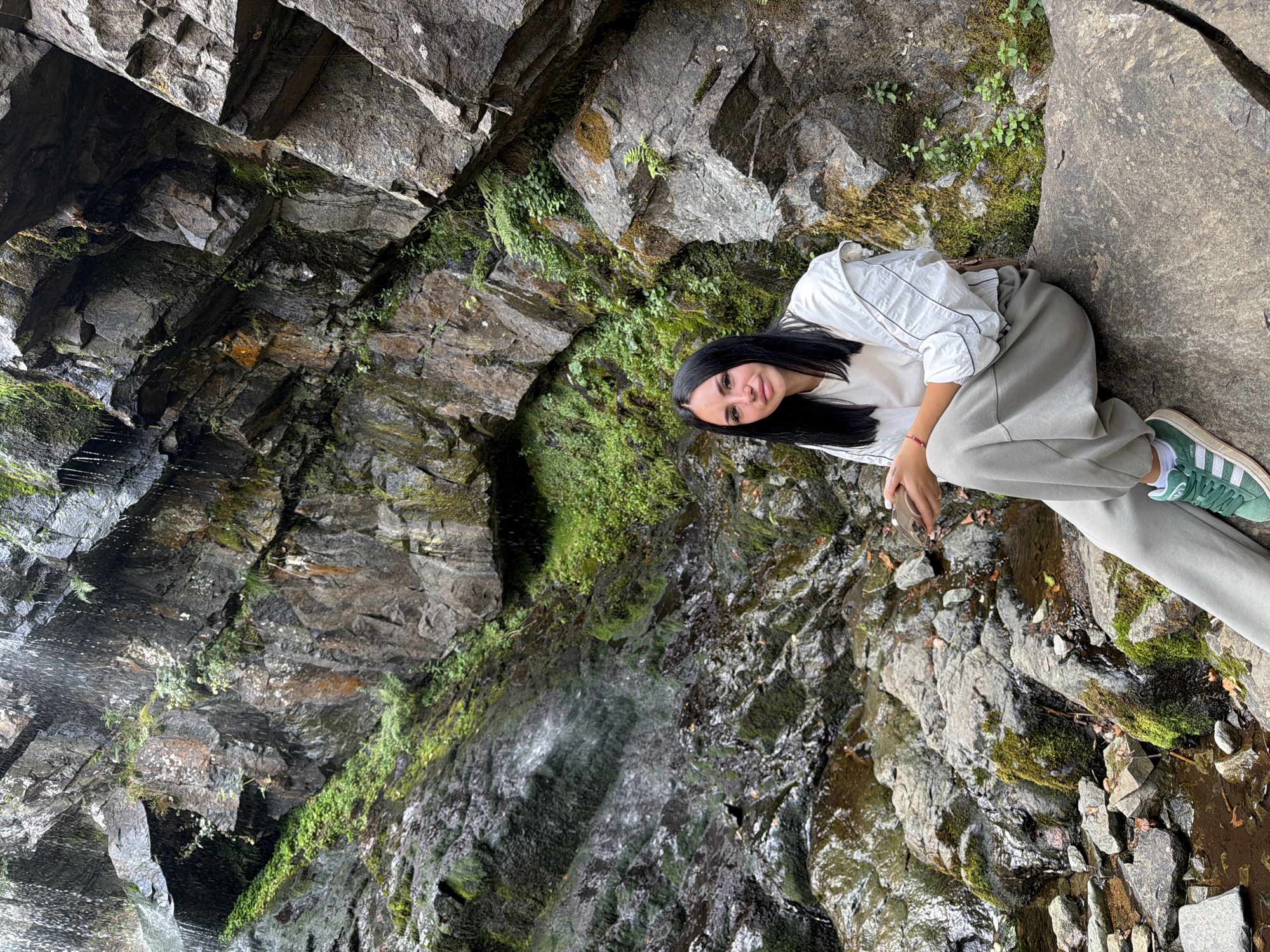 A young woman with long black hair and a light-colored outfit sits on a large rock in front of a rocky, moss-covered cliff face where a waterfall cascades down. She is holding a mobile phone in her hands. The surrounding environment is natural and rugged, with water, rocks, and vegetation. The image is a full-body shot of the woman and the natural surroundings. The location is the waterfall near Kopitoto on Vitosha Mountain, overlooking Sofia, Bulgaria.