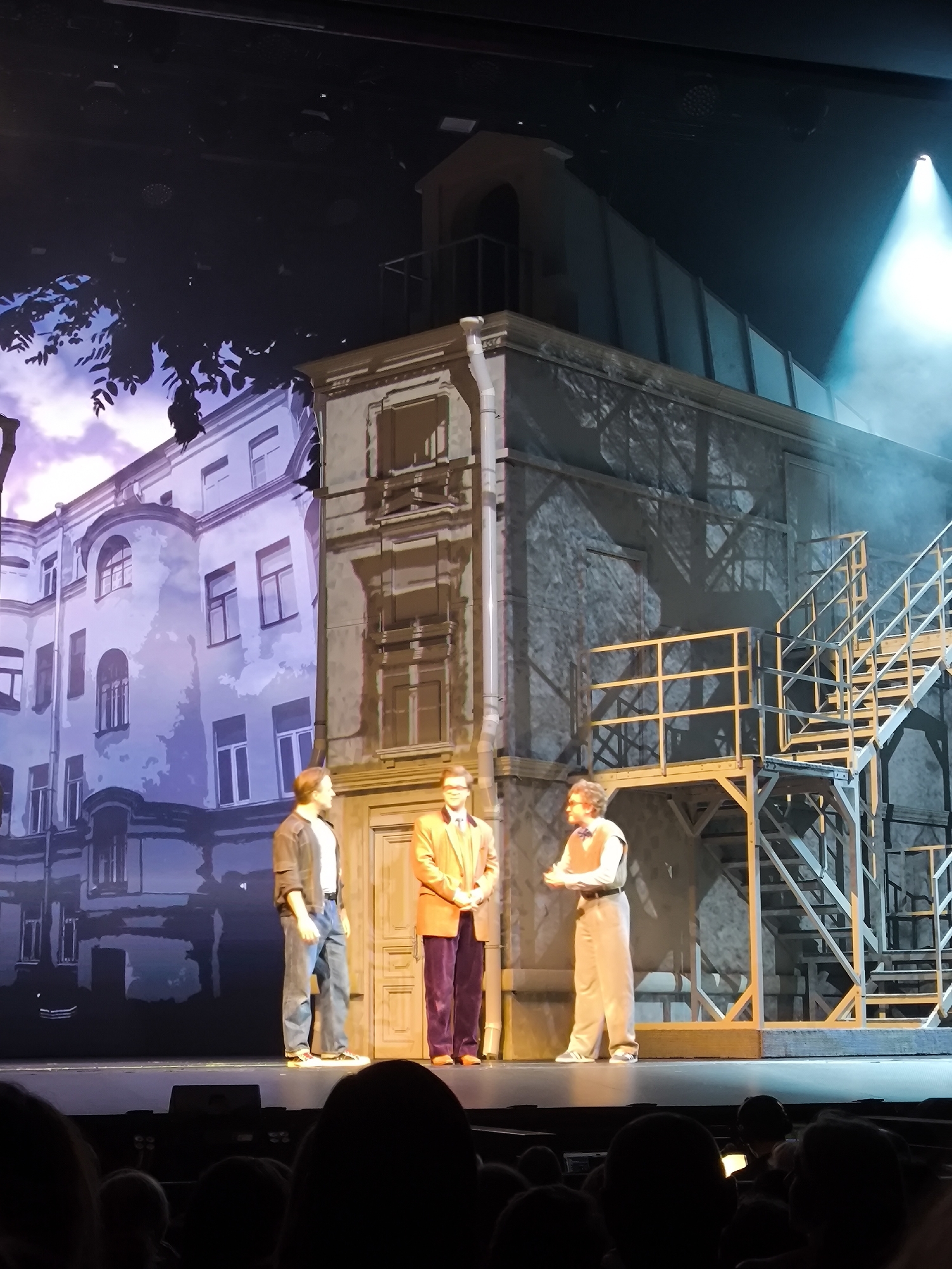 Three men stand on a stage set depicting a building facade and an external staircase. The man on the left wears a dark jacket, white t-shirt, and blue jeans. The man in the center wears an orange jacket, purple pants, and a white shirt. The man on the right wears a light-colored vest over a shirt and light-colored pants. The background features a projected image of a building with purple and blue tones and a dark, shadowy tree. A bright blue spotlight illuminates the top right of the stage. The stage floor is visible in the foreground, and the dark silhouettes of an audience are in the bottom of the frame.