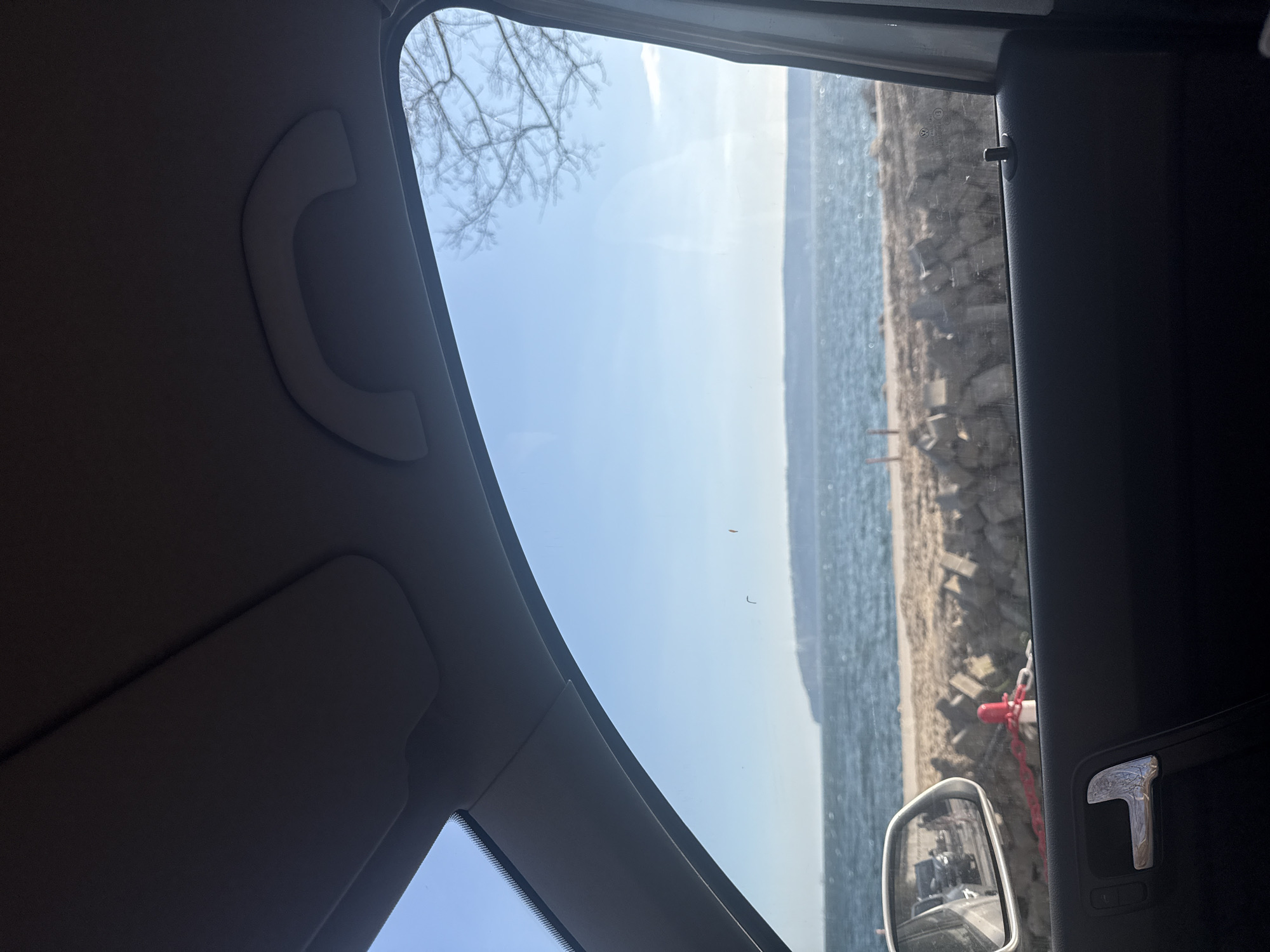 The image shows a view from inside a vehicle, likely a car, looking out towards a body of water and a coastline. The water appears calm and blue. The coastline consists of rocks and beach, with some man-made structures or barriers visible. A bare tree is visible through the front windshield. Within the vehicle, parts of the interior are visible, including a roof handle and the door panel. There is a side mirror on the door with a reflection. Based on the coastline and the presence of man-made structures, it's likely that the location is along the coast of Varna, Bulgaria. The specific section of the Varna coast cannot be pinpointed without more details. No people can be identified in the scene.