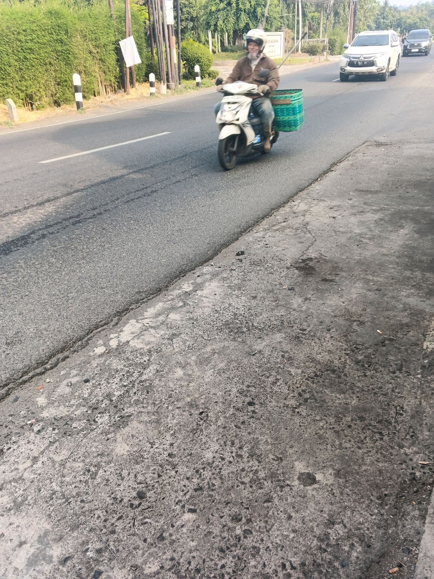 A man wearing a helmet and a brown jacket rides a white scooter down a road in Ungaran, Indonesia. The scooter has a large, green woven basket attached to the rear. The man appears to be looking forward and driving. Two cars are visible in the background, one white and one black. The road is paved and appears to be in good condition. There is a sidewalk to the right of the road, made of concrete. A dense green hedge lines the left side of the road, with several utility poles in front of it. The overall scene suggests a typical street view in a tropical or subtropical region.